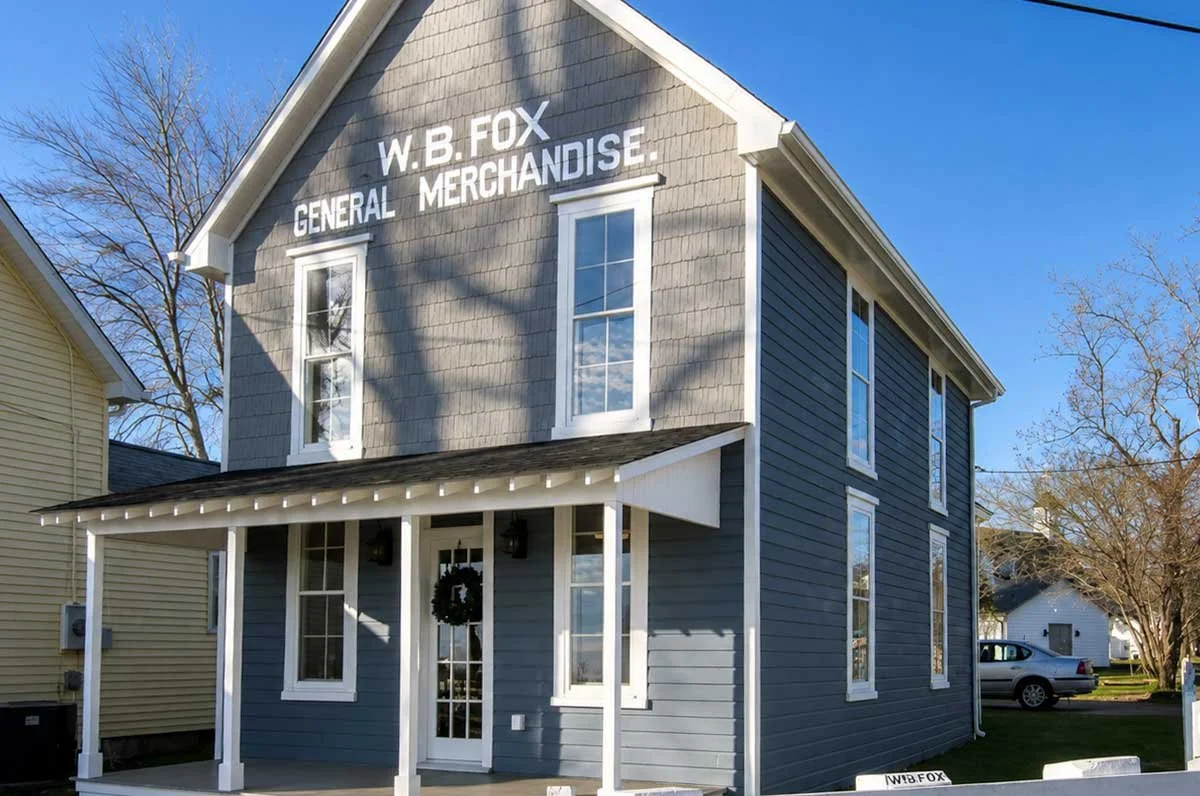A two-story building with gray siding and white trim, sign that reads 'W.B. Fox General Merchandise', front porch with a wreath on the door, and several windows on each floor. The sky is clear and trees are in the background.