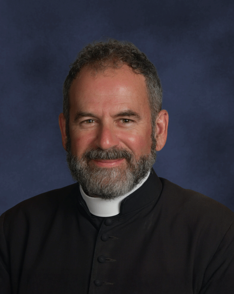 Portrait of a middle-aged man with a beard, wearing a black clerical shirt with a white clerical collar, against a dark blue background.