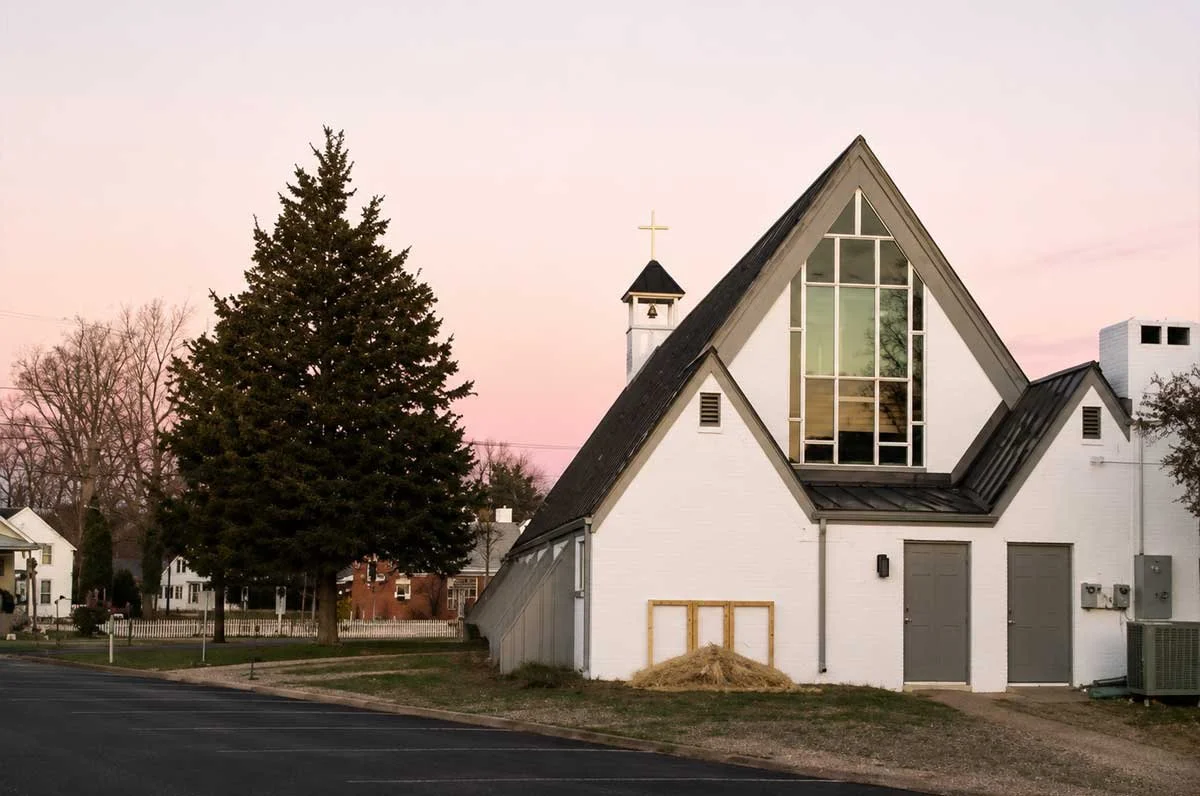 White church building with large triangular windows, a small bell tower topped with a cross, and a large evergreen tree in the background at sunset.