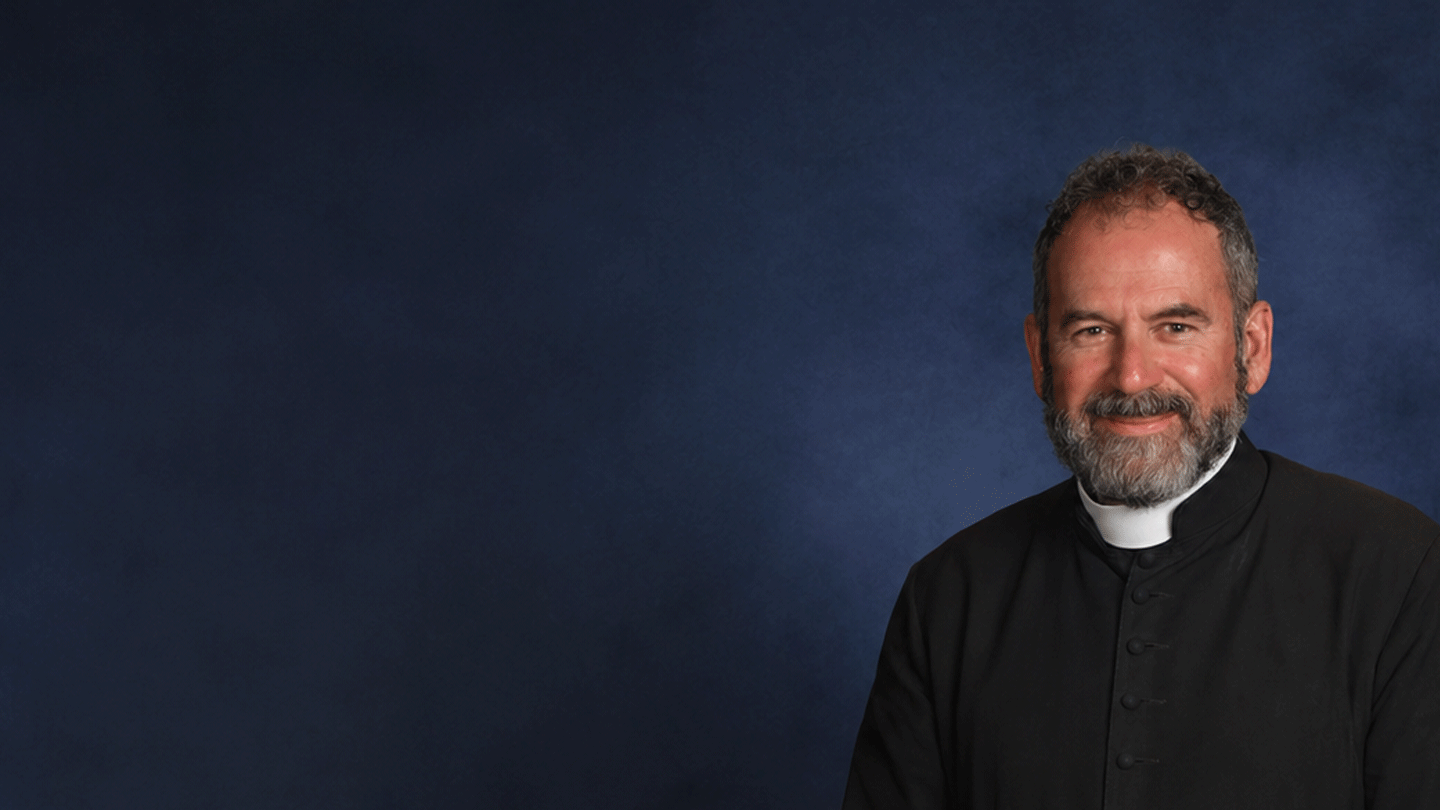A smiling man dressed as a priest with a beard, wearing a black clerical shirt with a white collar, against a dark blue background.