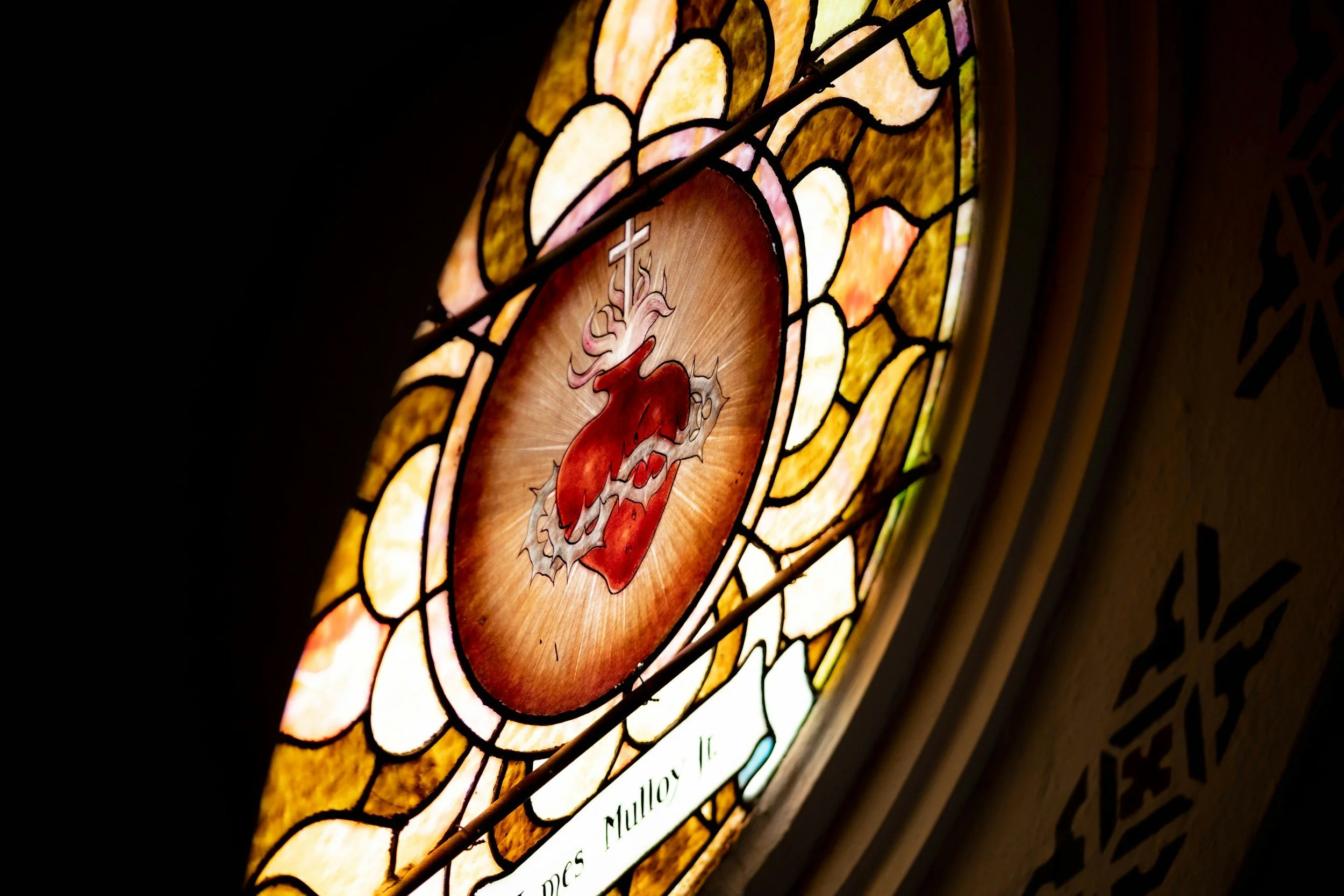 Close-up of a stained glass window depicting a red heart with a cross above it, surrounded by intricate colorful glass patterns.