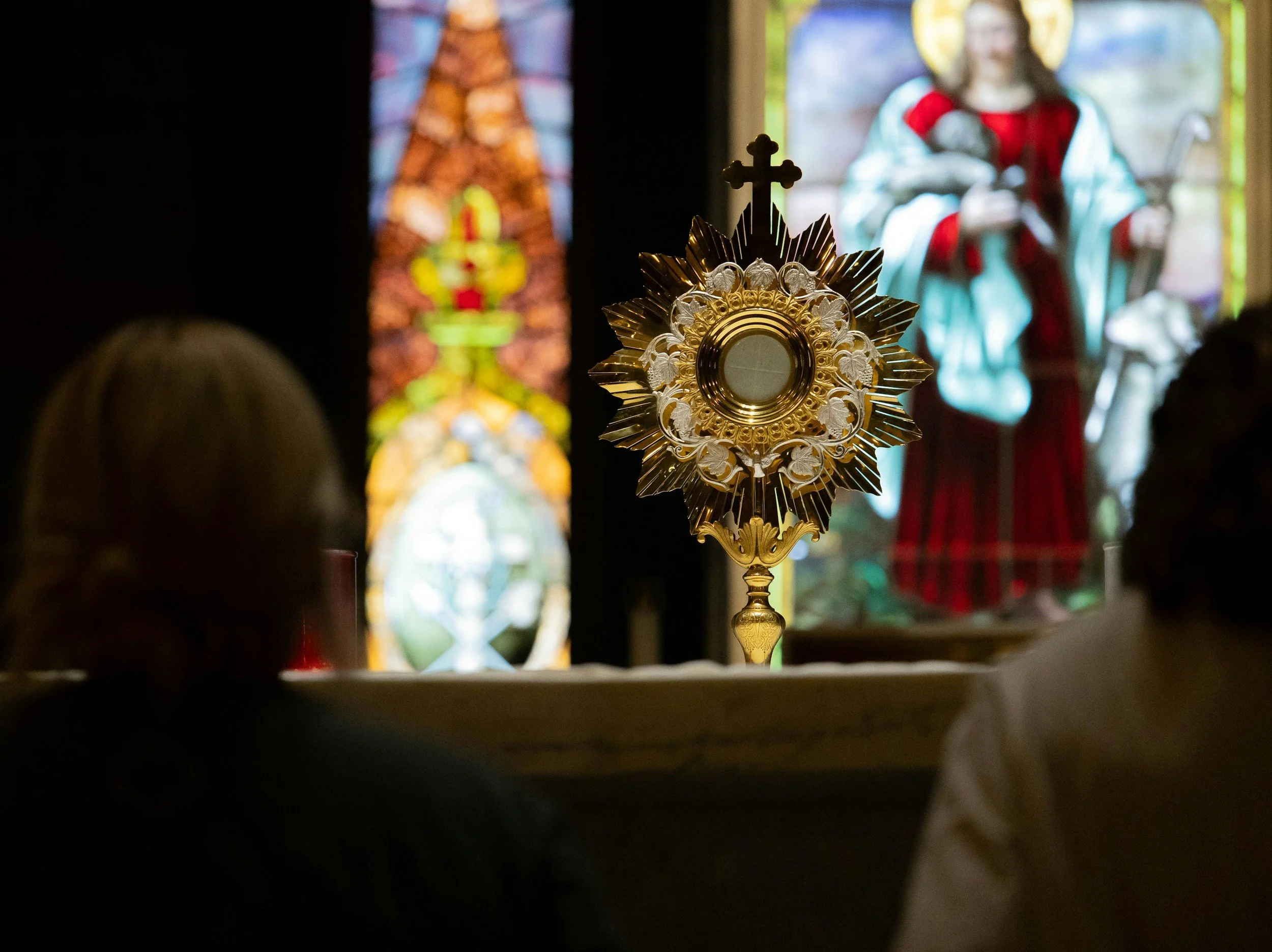 A religious monstrance in the foreground with stained glass images of saints and religious figures in the background.