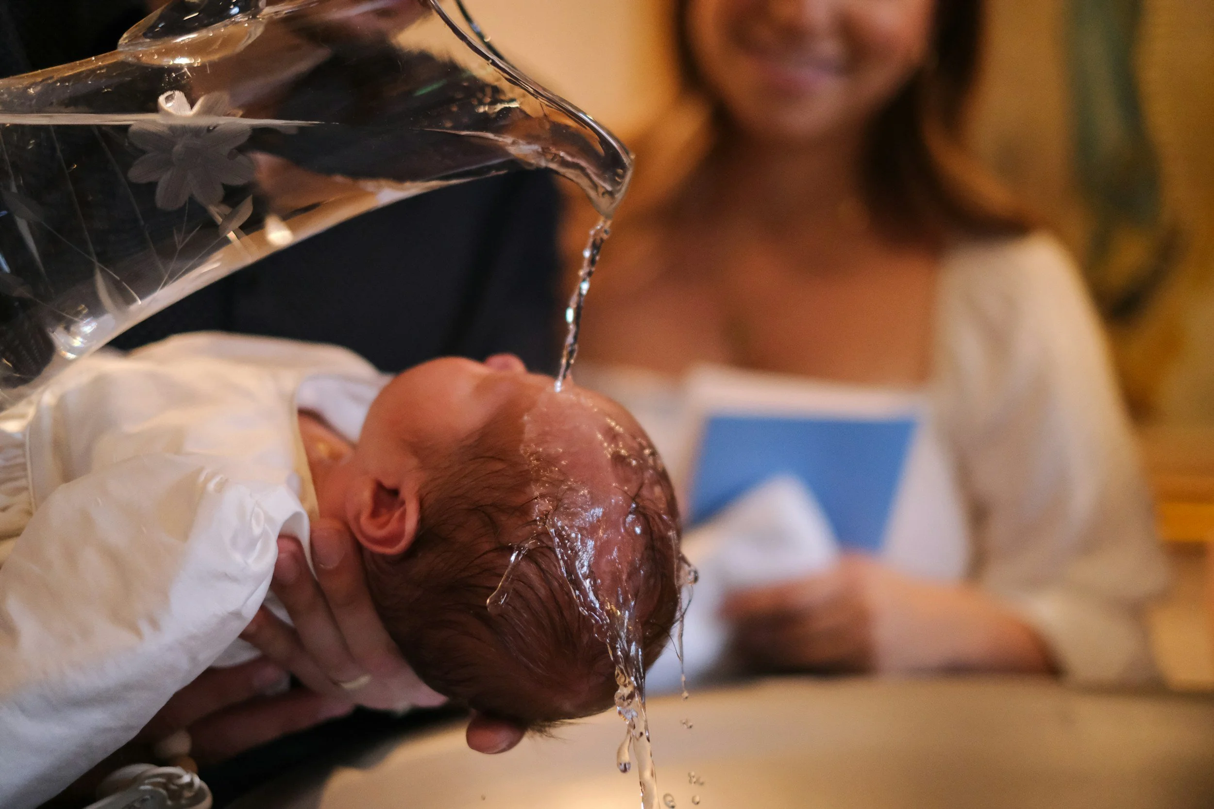 A person has their newborn baby's head gently supported while water is poured over the baby's head during a baptism or christening ceremony.