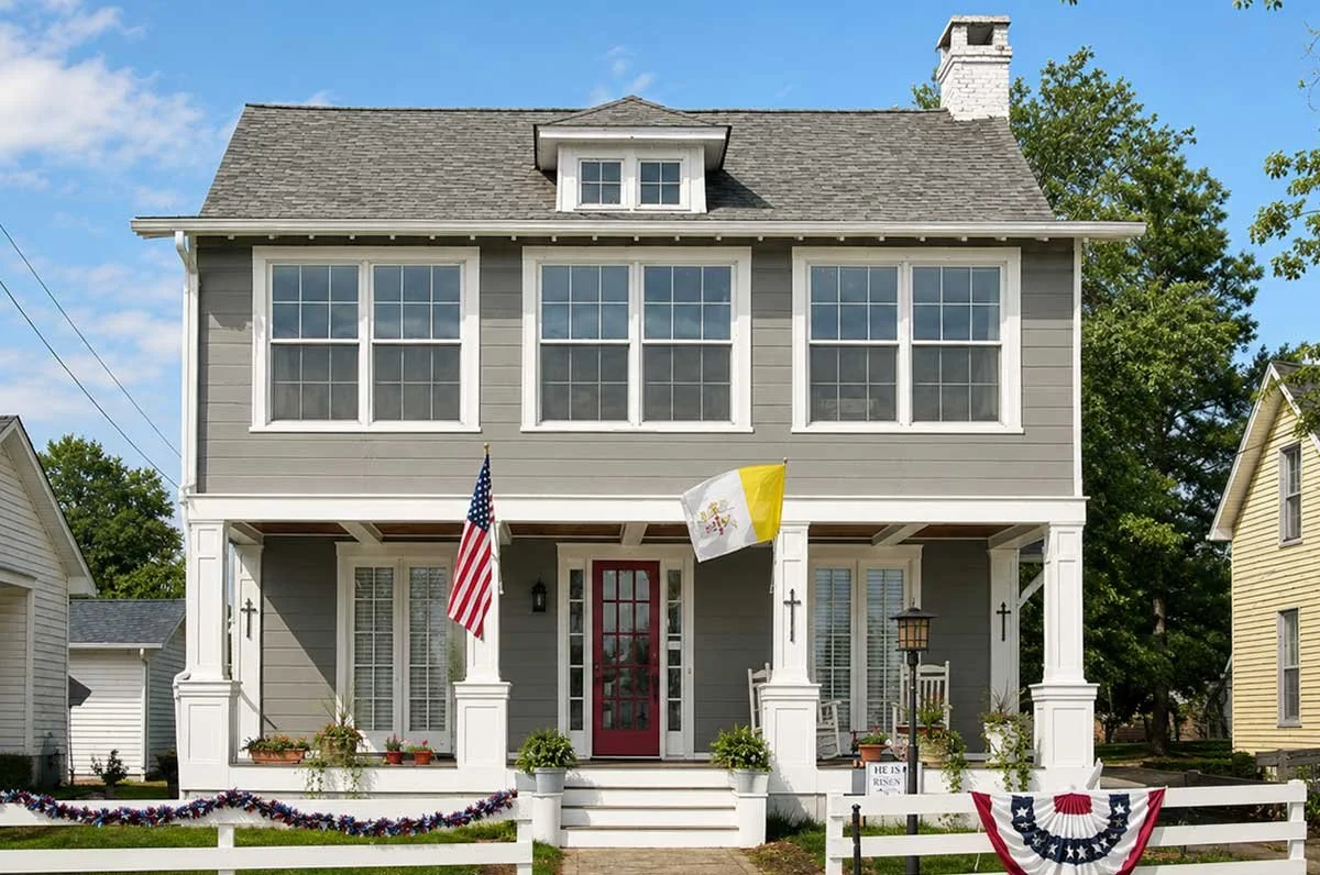 A two-story house decorated for a patriotic celebration with American flags, a flag with yellow and white stripes, and red, white, and blue bunting on the front porch, with a white picket fence in front.