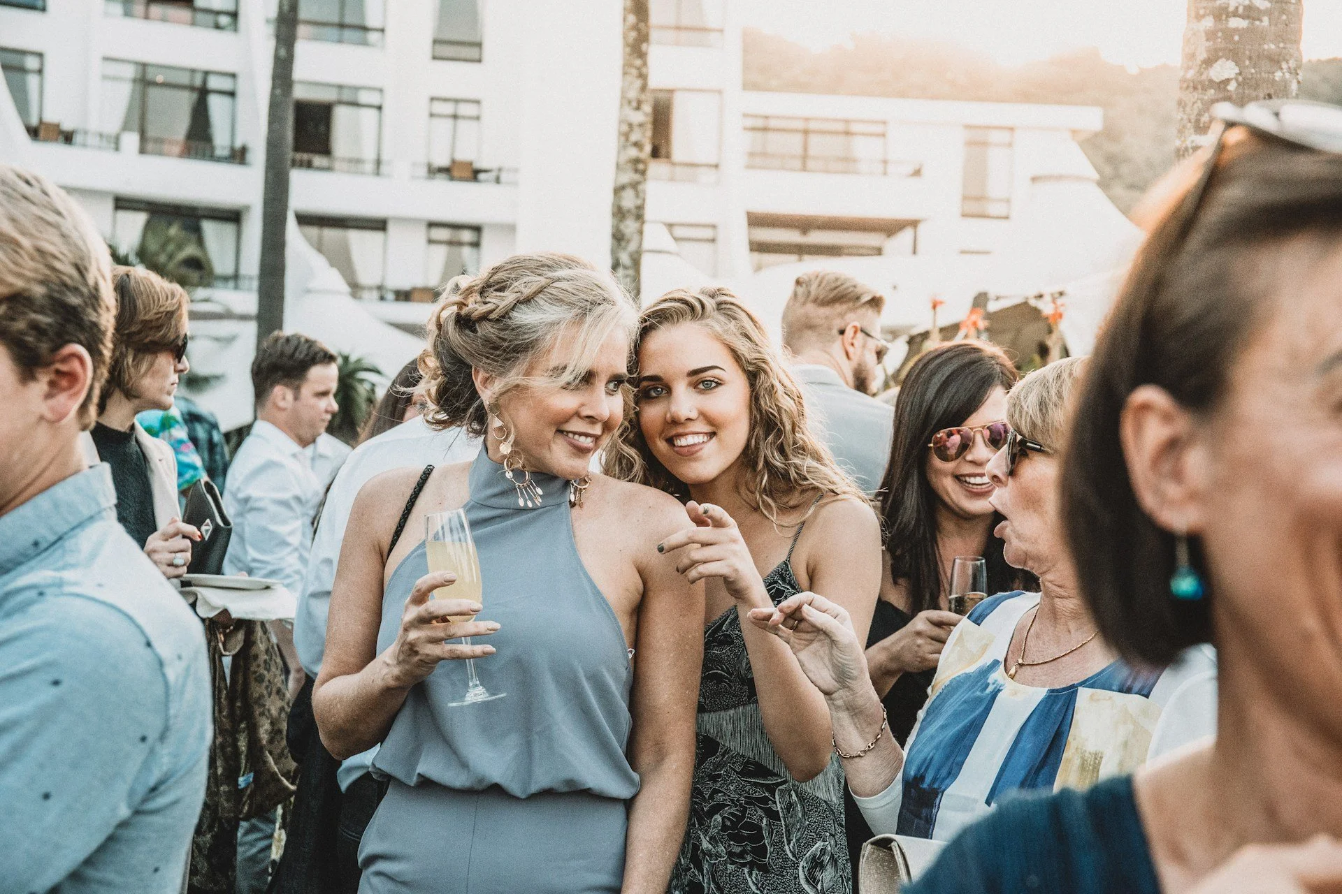 A group of people at an outdoor event, with two women in the center smiling and holding drinks, surrounded by other attendees, with modern buildings in the background during sunset.