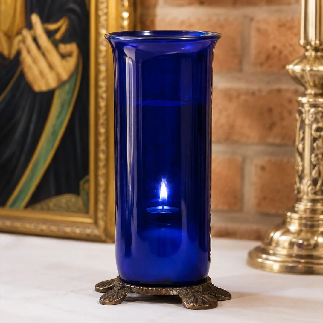 A tall, cobalt blue glass vase with a candle inside, placed on a decorative stand in front of a framed painting and a brass candlestick, on a white surface.