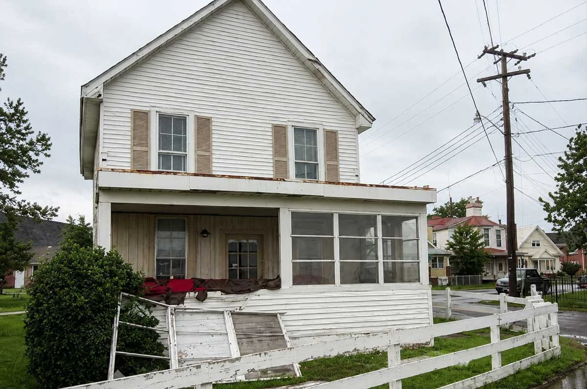 An old, two-story house with peeling white paint and broken railing in the front yard, with utility poles and other houses in the background on a cloudy day.