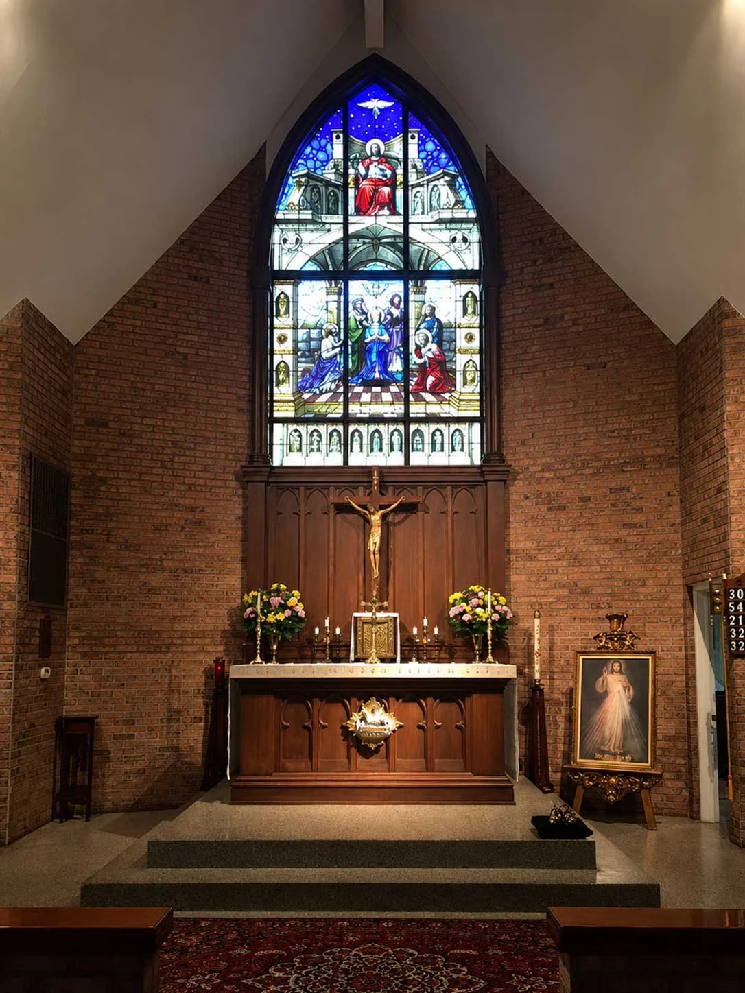Inside a church, there is a wooden altar with a crucifix, flowers, candles, and religious symbols. Behind the altar is a large stained glass window depicting a religious scene with figures, set against brick walls.