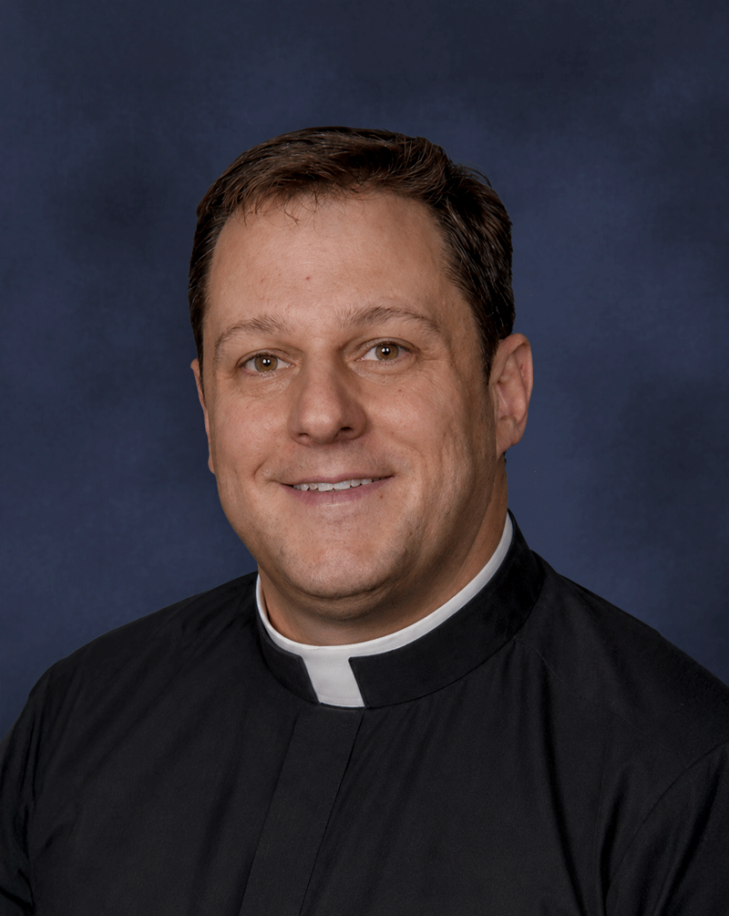 Portrait of a man in clergy attire with a clerical collar, smiling against a dark blue background.