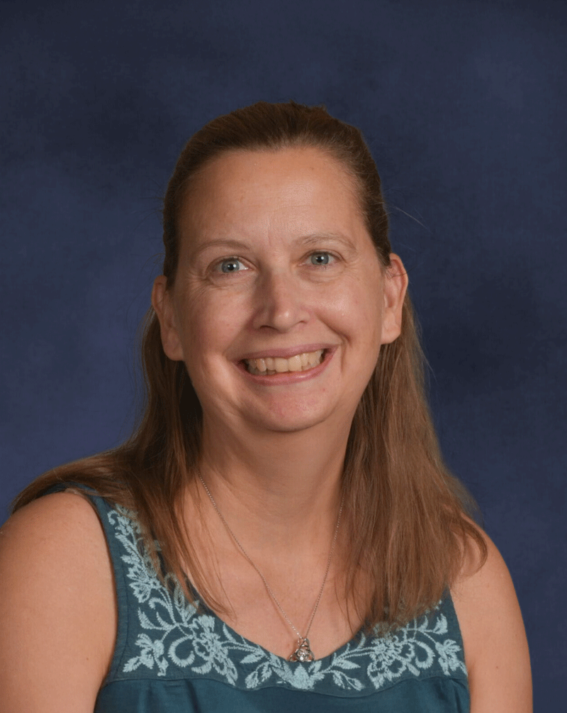 A woman with long light brown hair in a ponytail smiling at the camera, wearing a blue sleeveless top with white embroidery and a silver necklace, against a dark blue background.