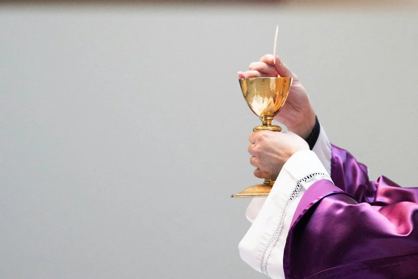 Person in purple liturgical robes holding a golden chalice with a white candle or wafer.