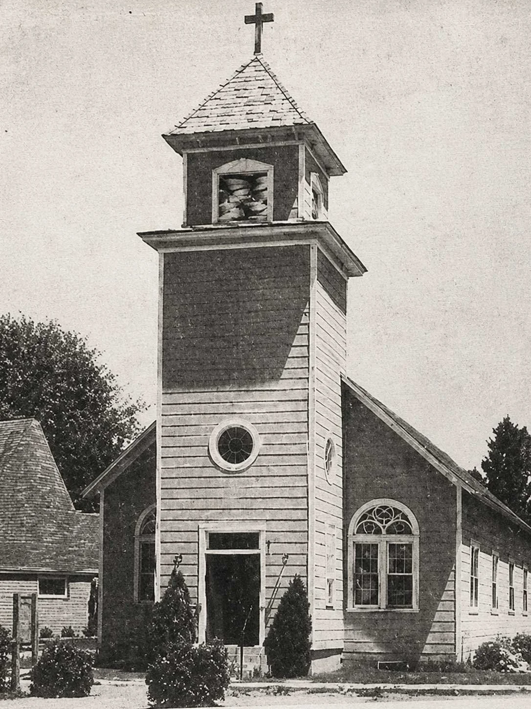 A black-and-white photo of an old wooden church with a bell tower topped by a cross, surrounded by small bushes and trees.