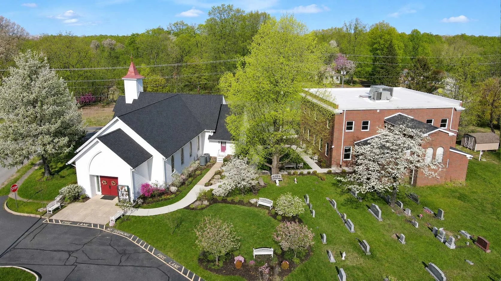 Aerial view of a church surrounded by blooming trees and a graveyard, with a parking lot nearby.