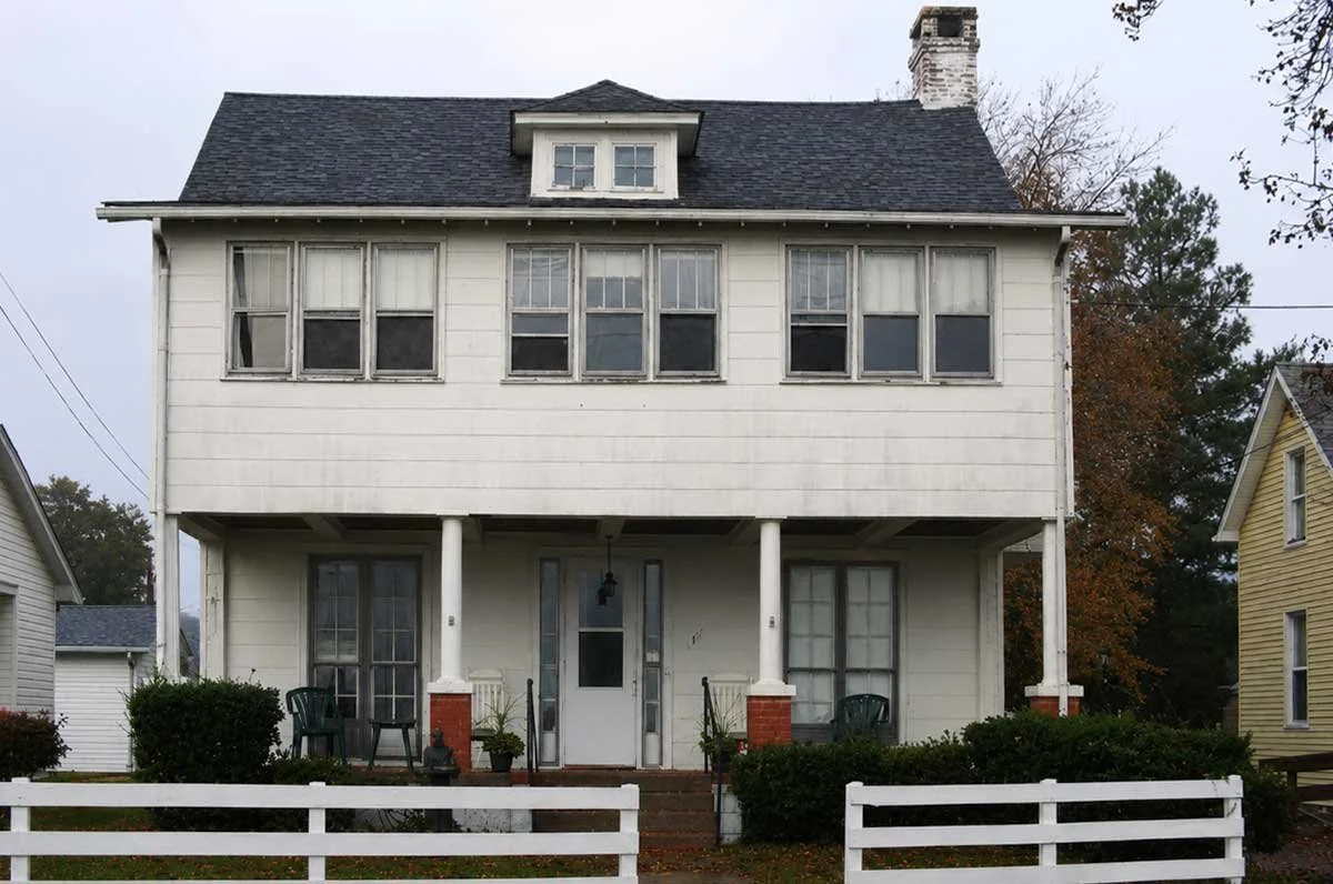 A white, three-story house with a black roof, front porch, and multiple windows.