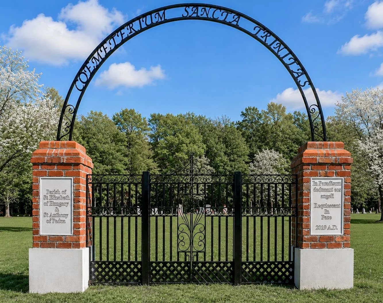 Entrance gate to a cemetery with brick pillars and metal gates, surrounded by trees and a blue sky.