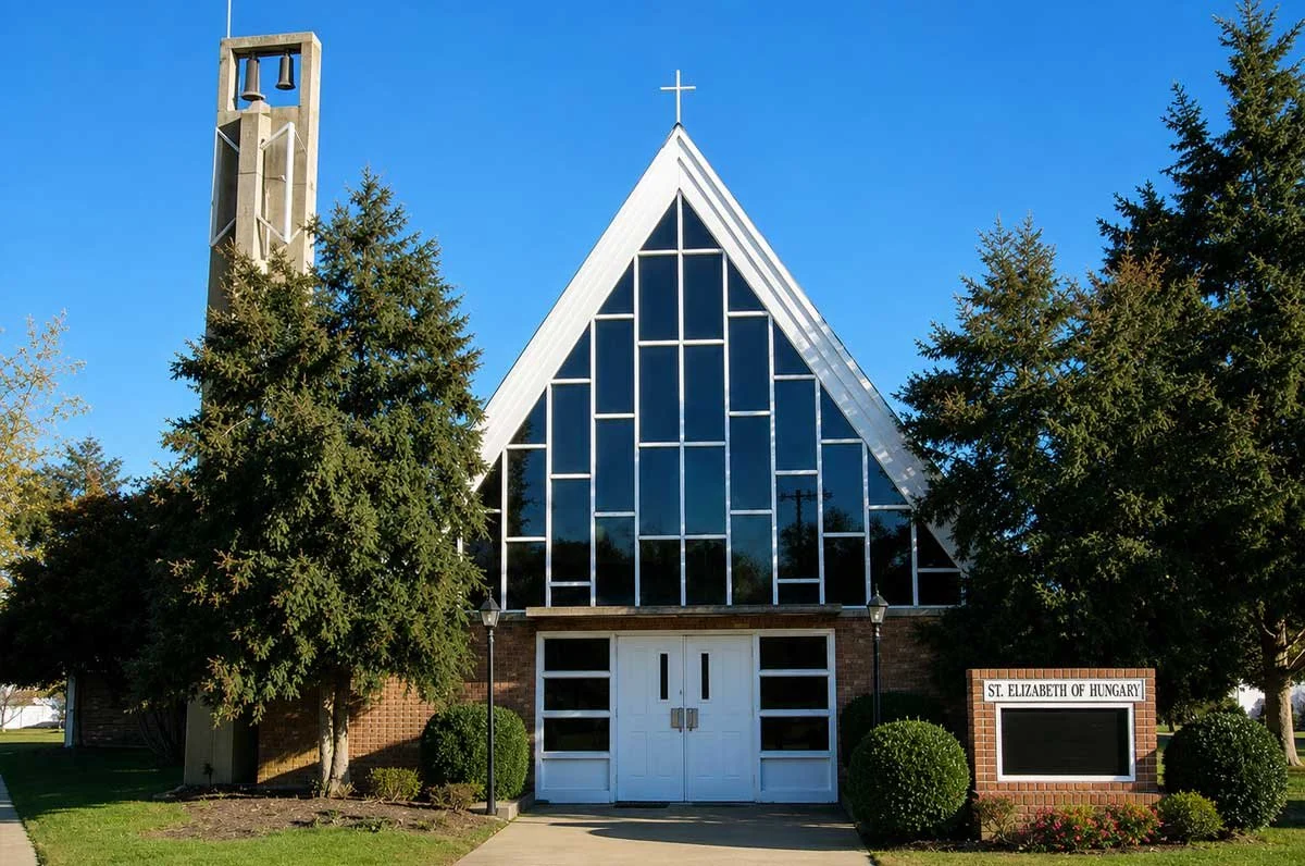 Front view of a church named St. Elizabeth of Hungary, with a triangular glass facade, a bell tower on the left, and surrounding trees and bushes on a clear day.