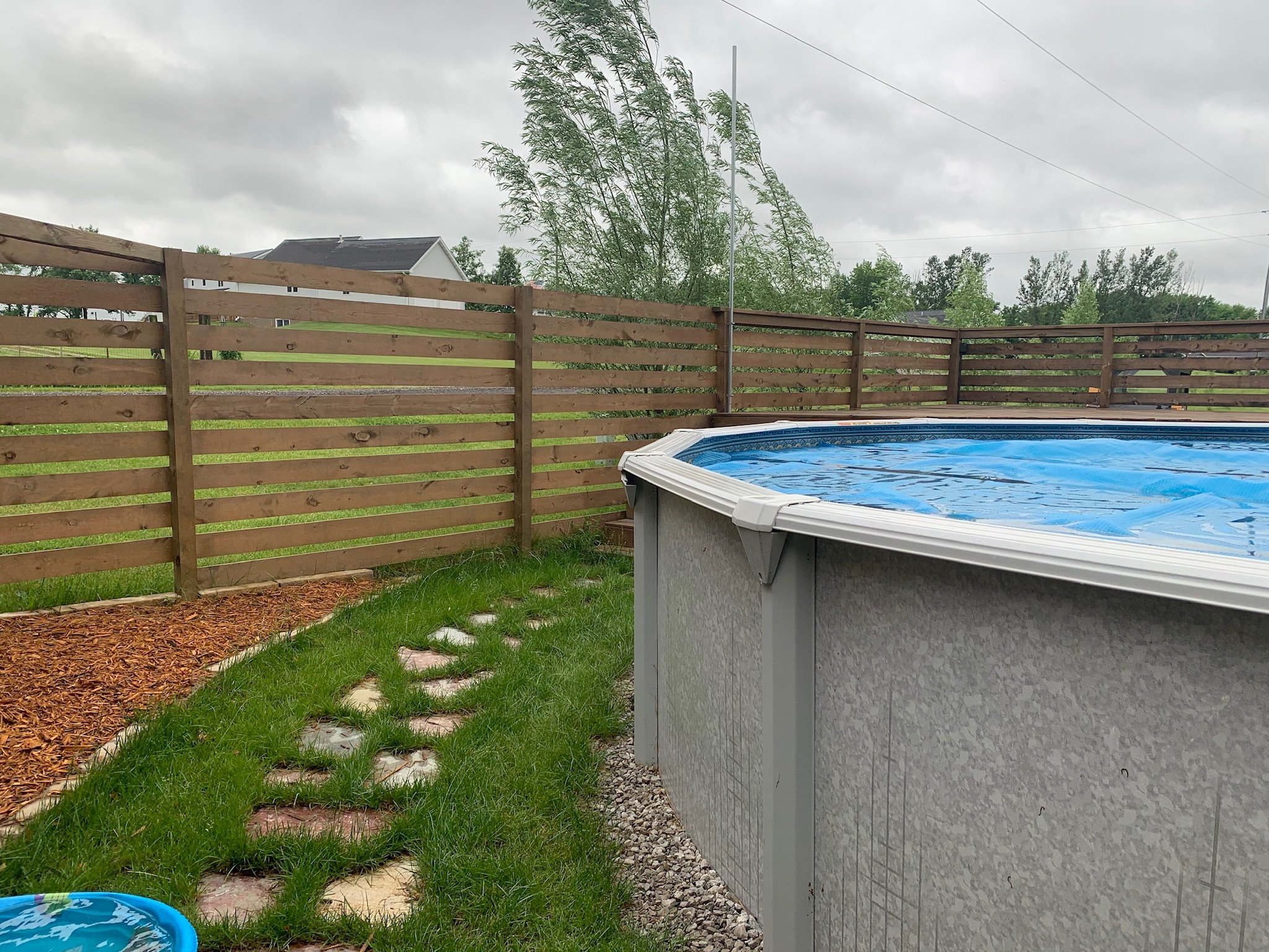 A backyard scene with a wooden fence, an above ground pool, green grass, and a cloudy sky. Iowa City. Stecker Home Services. Home Renovations. Iowa City Contractor. 