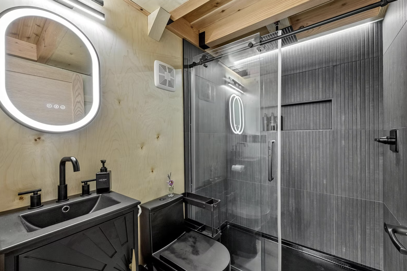Modern bathroom with a black vanity, circular mirror, black faucet, black toilet, and a glass shower enclosure with gray tile walls and a heated towel rack. Iowa City. Stecker Home Services. Home Renovations. Iowa City Contractor. 