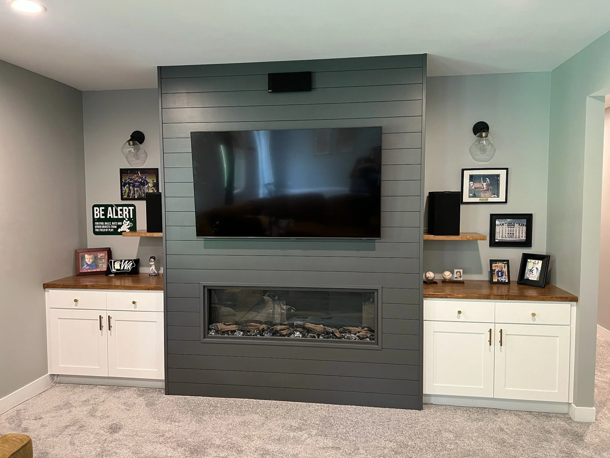 Living room with a large flat-screen TV mounted on a gray wooden feature wall, flanked by two wall sconces with glass shades. Below the TV are built-in cabinets with white doors and brass handles. Iowa City. Stecker Home Services. Home Renovations