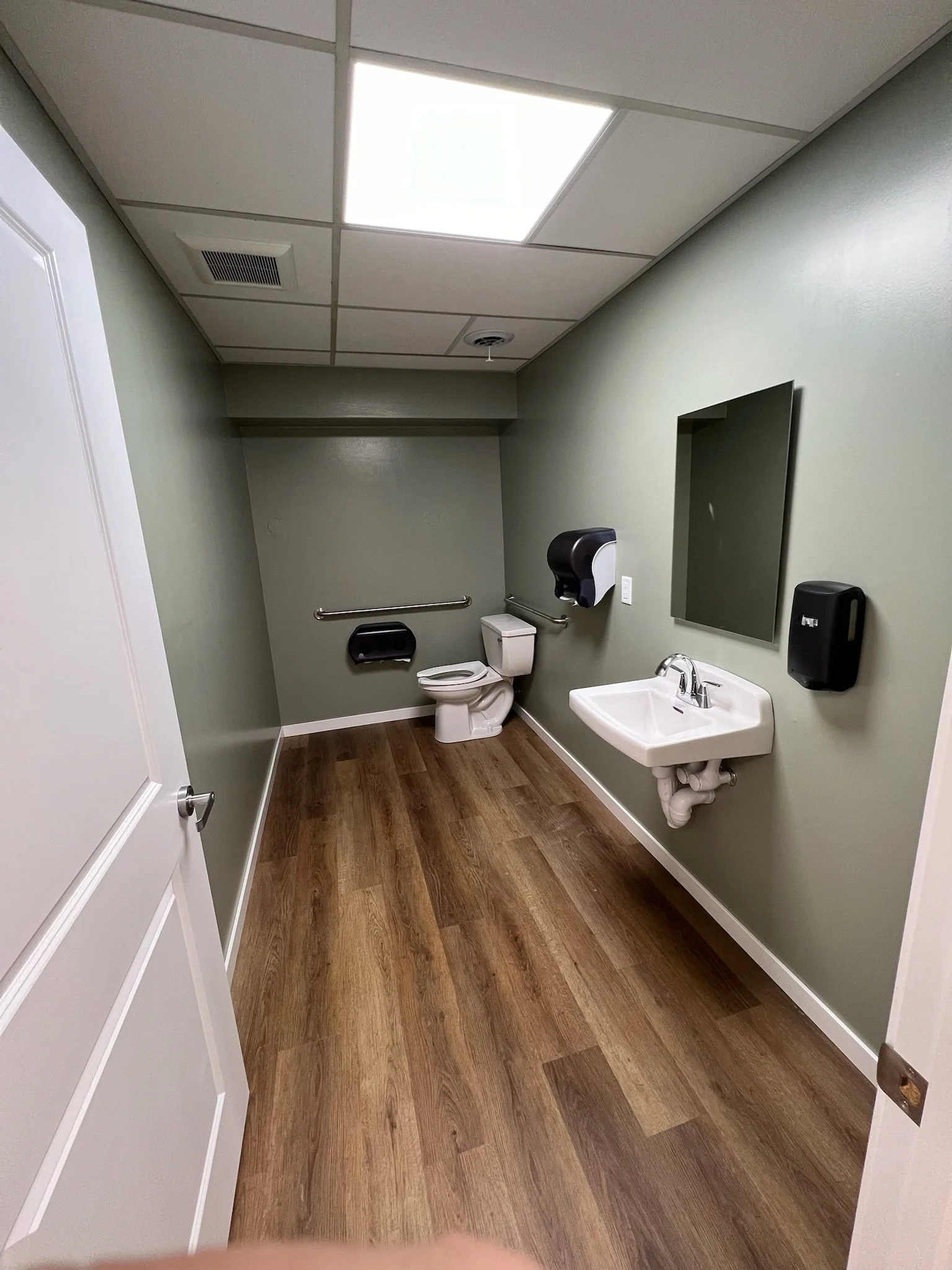 A clean and empty public restroom with green walls, a white toilet, a sink, a large mirror, a paper towel dispenser, and a soap dispenser. The ceiling has fluorescent lighting and an air vent.