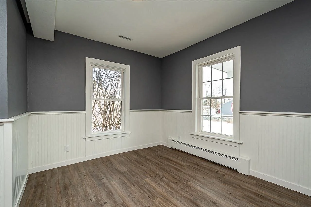 Empty room with gray upper walls, white beadboard wainscoting, two windows with view of trees and snow, brown wood floor, white baseboard heater. Iowa City. Stecker Home Services. Home Renovations. Iowa City Contractor. 