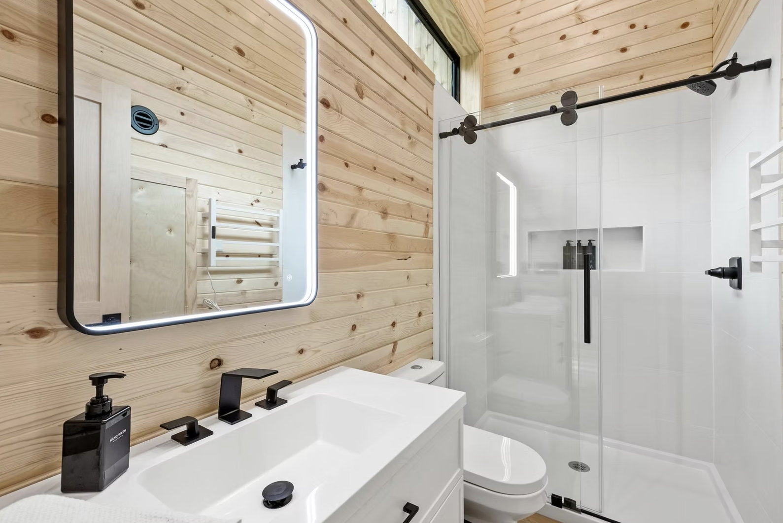 Modern bathroom with wooden wall, illuminated mirror, black fixtures, shower with sliding glass door, and white walls and fixtures. Iowa City. Stecker Home Services. Home Renovations. Iowa City Contractor. 