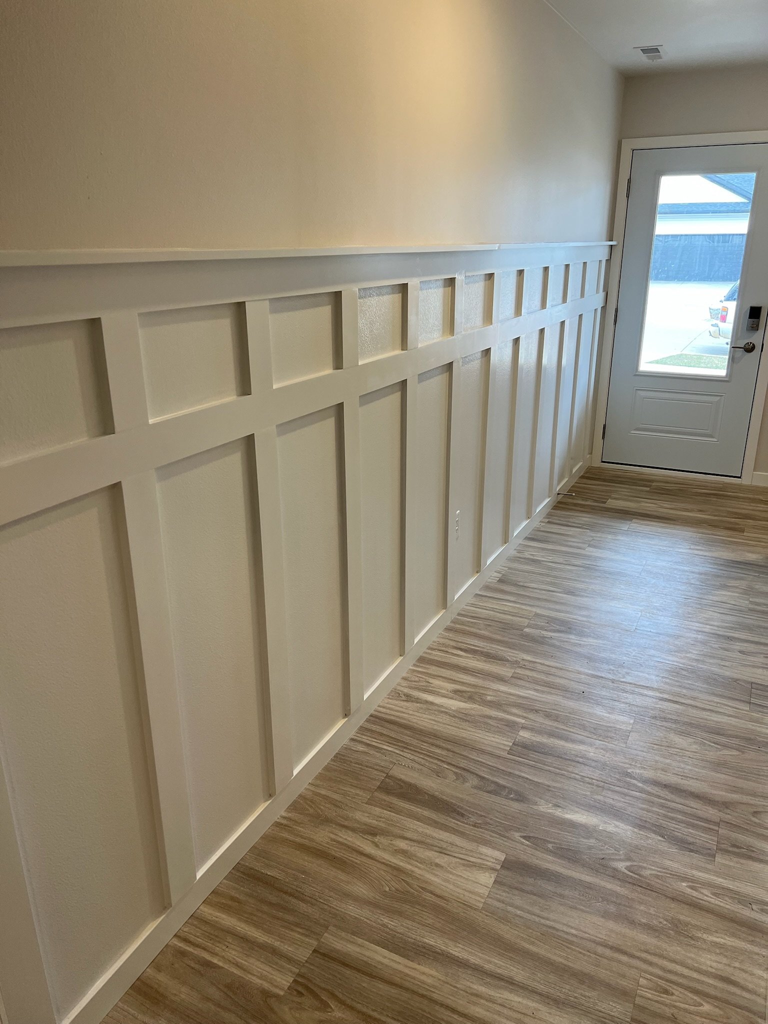 Interior hallway with wooden flooring, a white wall with decorative wood paneling, and a glass door leading outside, allowing natural light to enter. Iowa City. Stecker Home Services. Home Renovations. 