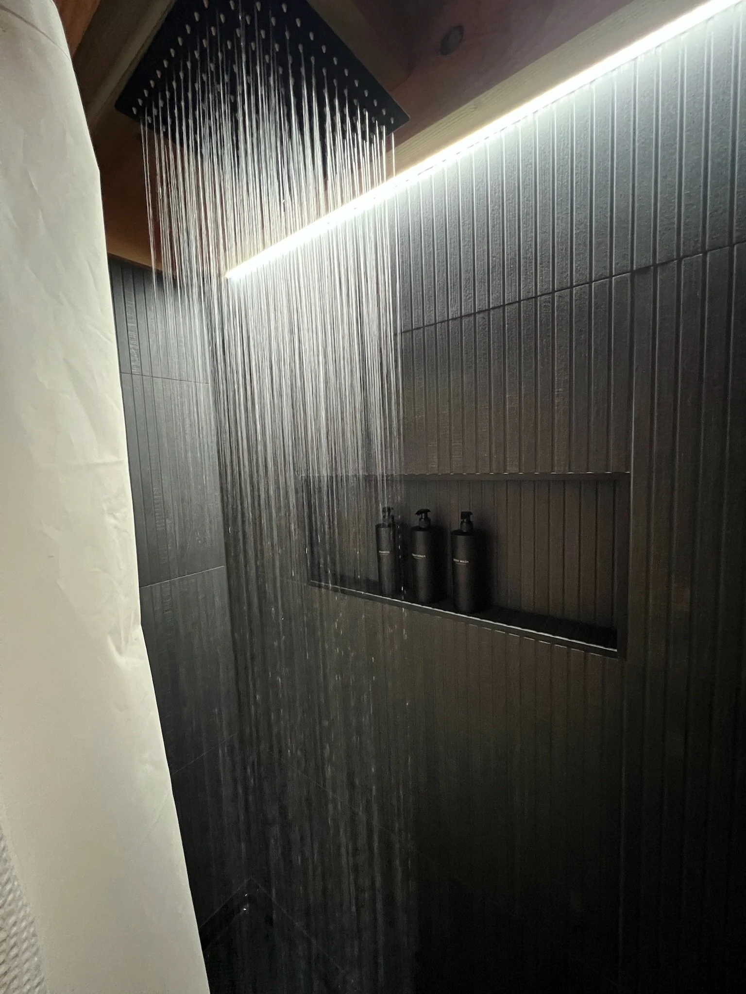 Shower with black square shower head, water flowing, gray tiled wall, and three black bottles on a ledge. Iowa City. Stecker Home Services. Home Renovations. Iowa City Contractor. 