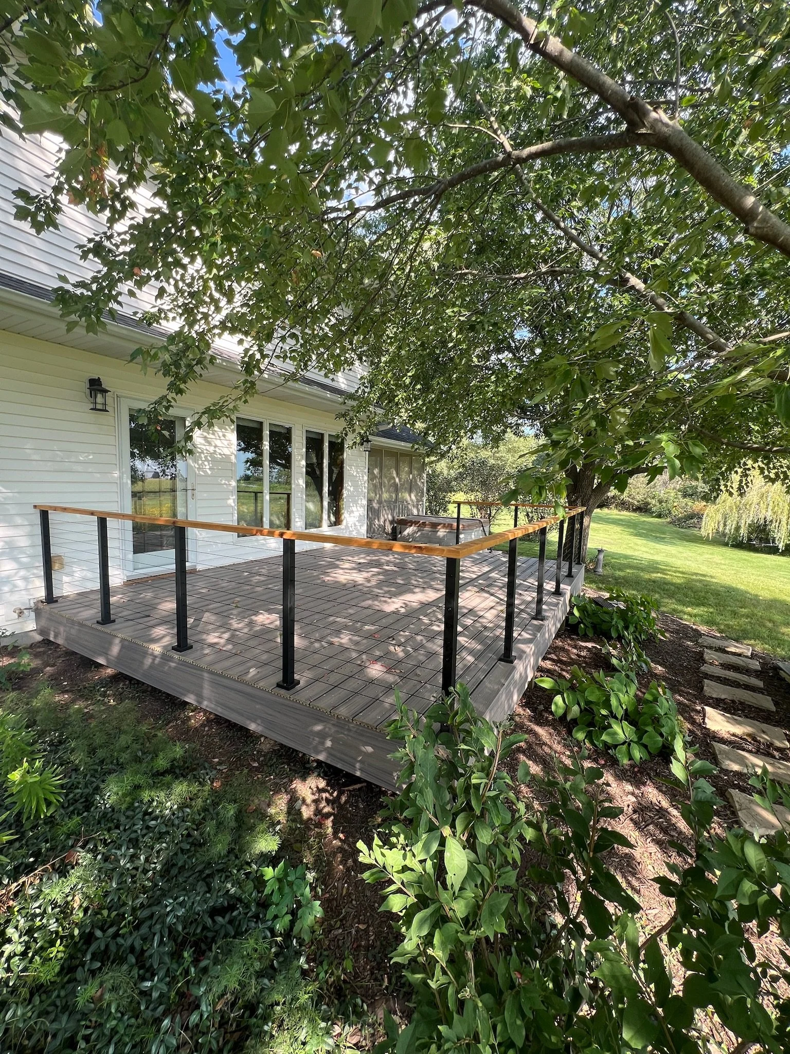 A backyard deck attached to a white house, surrounded by leafy trees and green plants, with a lawn in the background. Iowa City. Stecker Home Services. Home Renovations. Iowa City Contractor. 