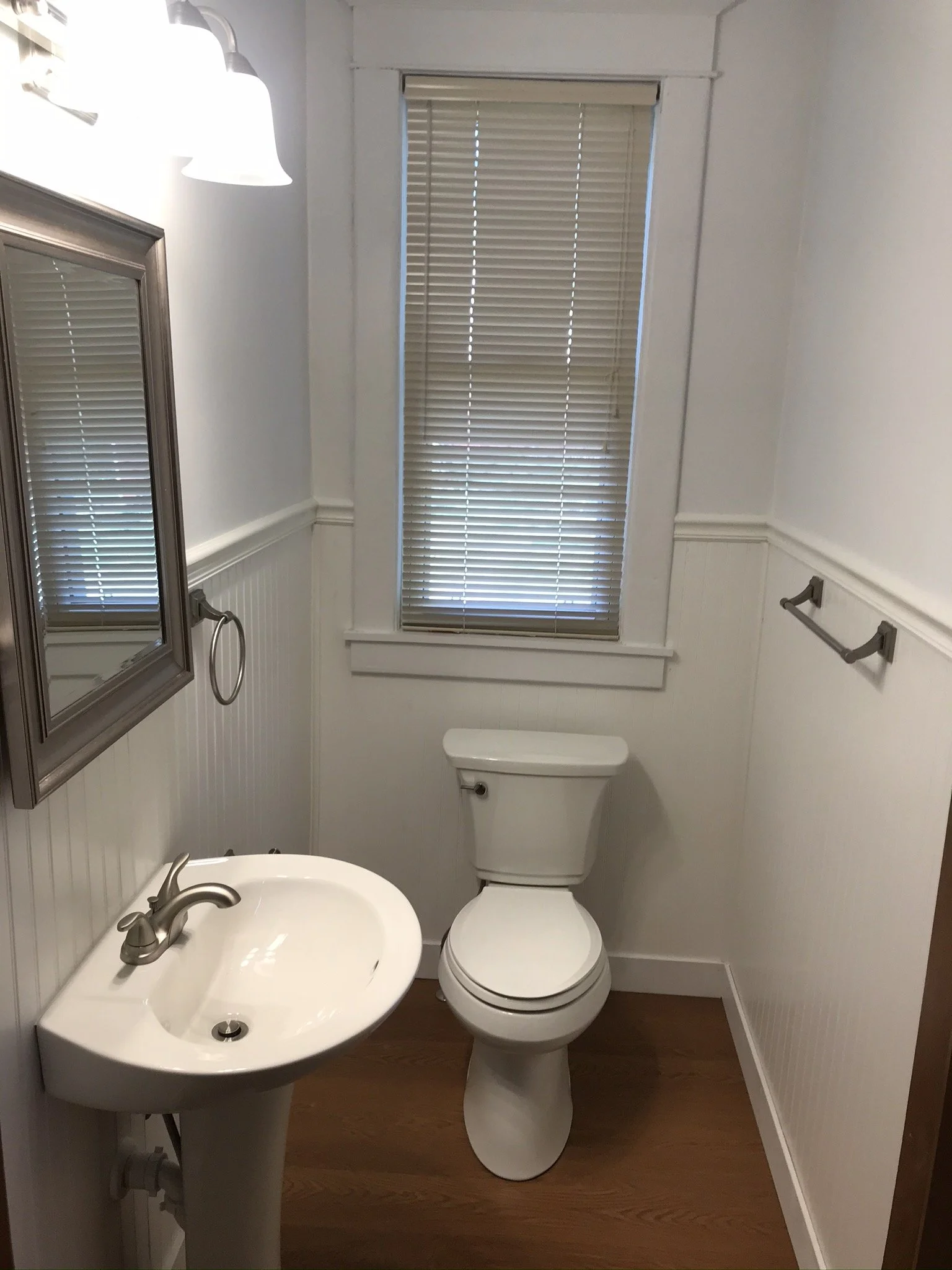 Small bathroom with white walls, a window with blinds, a white toilet, a small white sink, mirror, and a towel rack. Iowa City. Stecker Home Services. Home Renovations. Iowa City Contractor. 