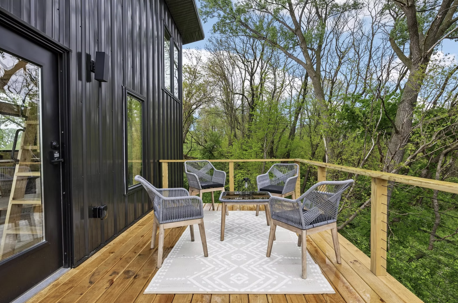 A black metal exterior building with a wooden balcony, four outdoor chairs around a glass table, and a view of trees and green foliage in the background. Iowa City. Stecker Home Services. Home Renovations. Iowa City Contractor. 