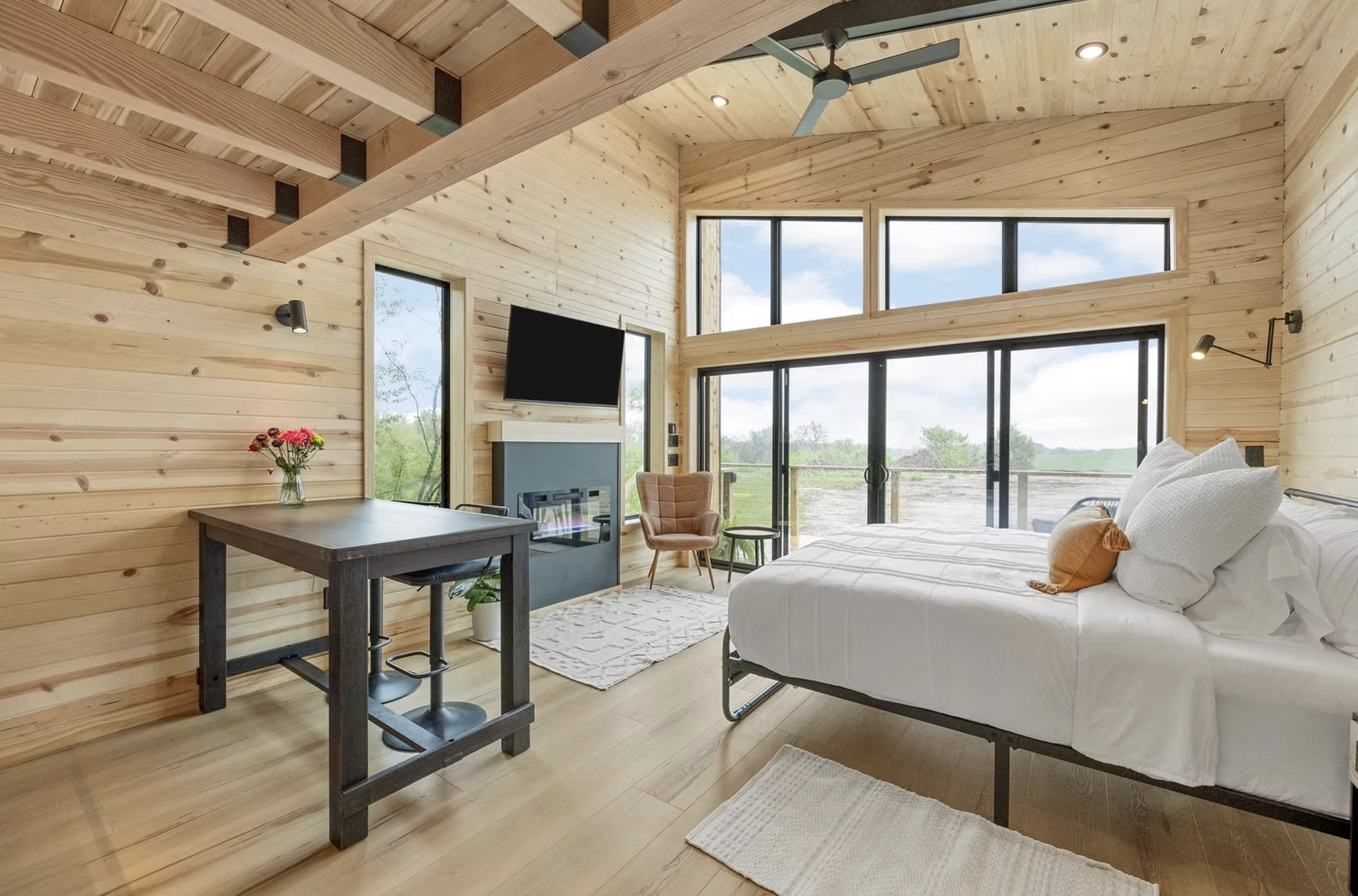 A modern bedroom with wooden walls and ceiling, a large bed with white linens, a table with a vase of flowers, a TV mounted above a fireplace, and large windows showing a green outdoor landscape. Iowa City. Stecker Home Services. Home Renovations.