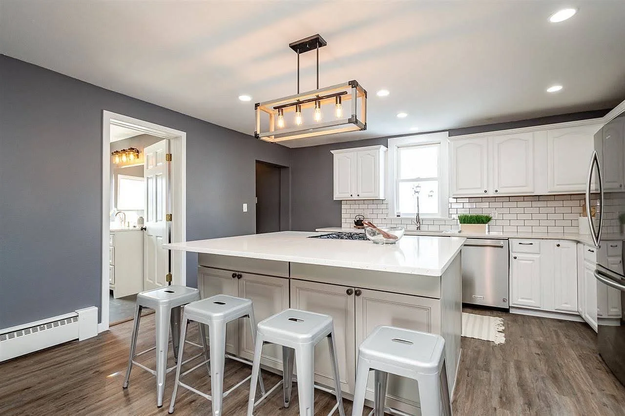Modern kitchen with white cabinets, gray wall, wooden flooring, and a kitchen island with four white stools. Iowa City. Stecker Home Services. Home Renovations. Iowa City Contractor. 