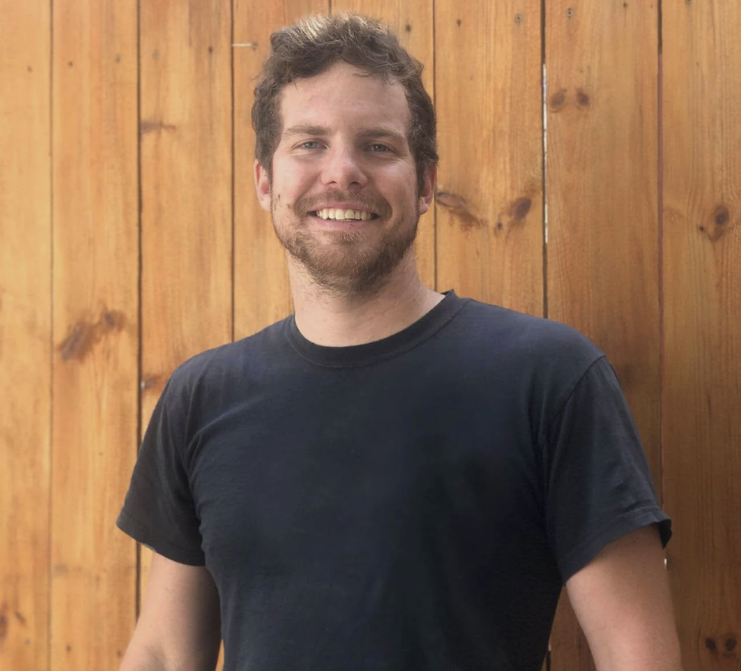 A smiling man with curly brown hair and a beard, wearing a black t-shirt, standing in front of a wooden fence. Iowa City. Stecker Home Services. Home Renovations.