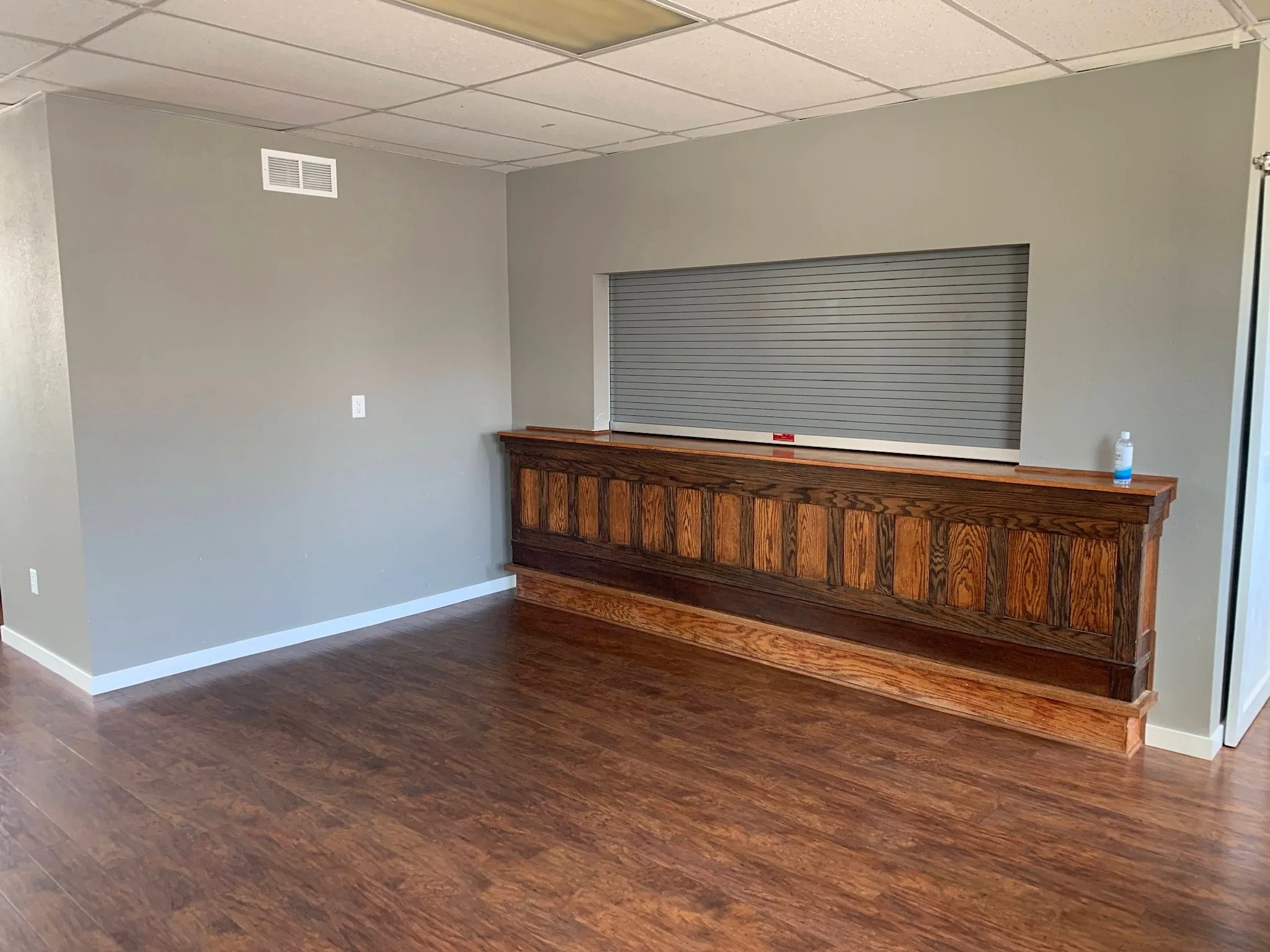 Empty interior room with gray walls, hardwood flooring, a window with a closed metal shutter, and a wooden counter below the window.  Iowa City. Stecker Home Services. Home Renovations. Iowa City Contractor. 