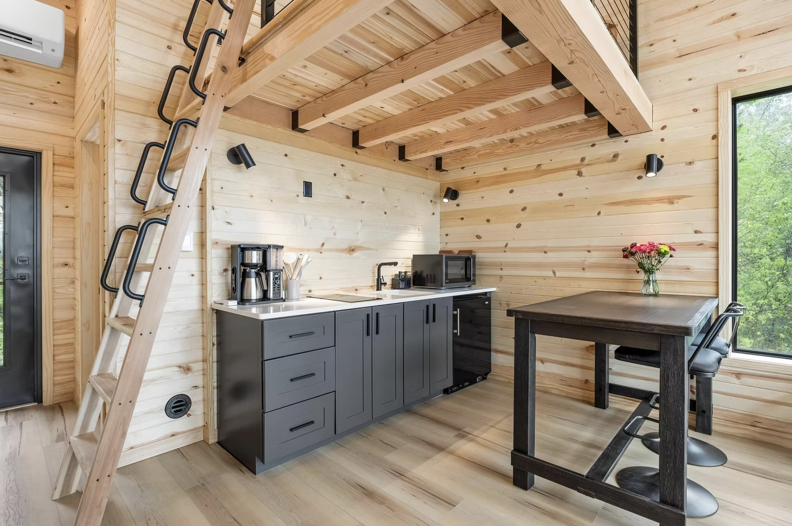 A cozy kitchen with wood-paneled walls and ceiling, a small kitchen counter, a black mini fridge, a dark wooden dining table with two chairs, and a window showing green trees outside. Iowa City. Stecker Home Services. Home Renovations. 
