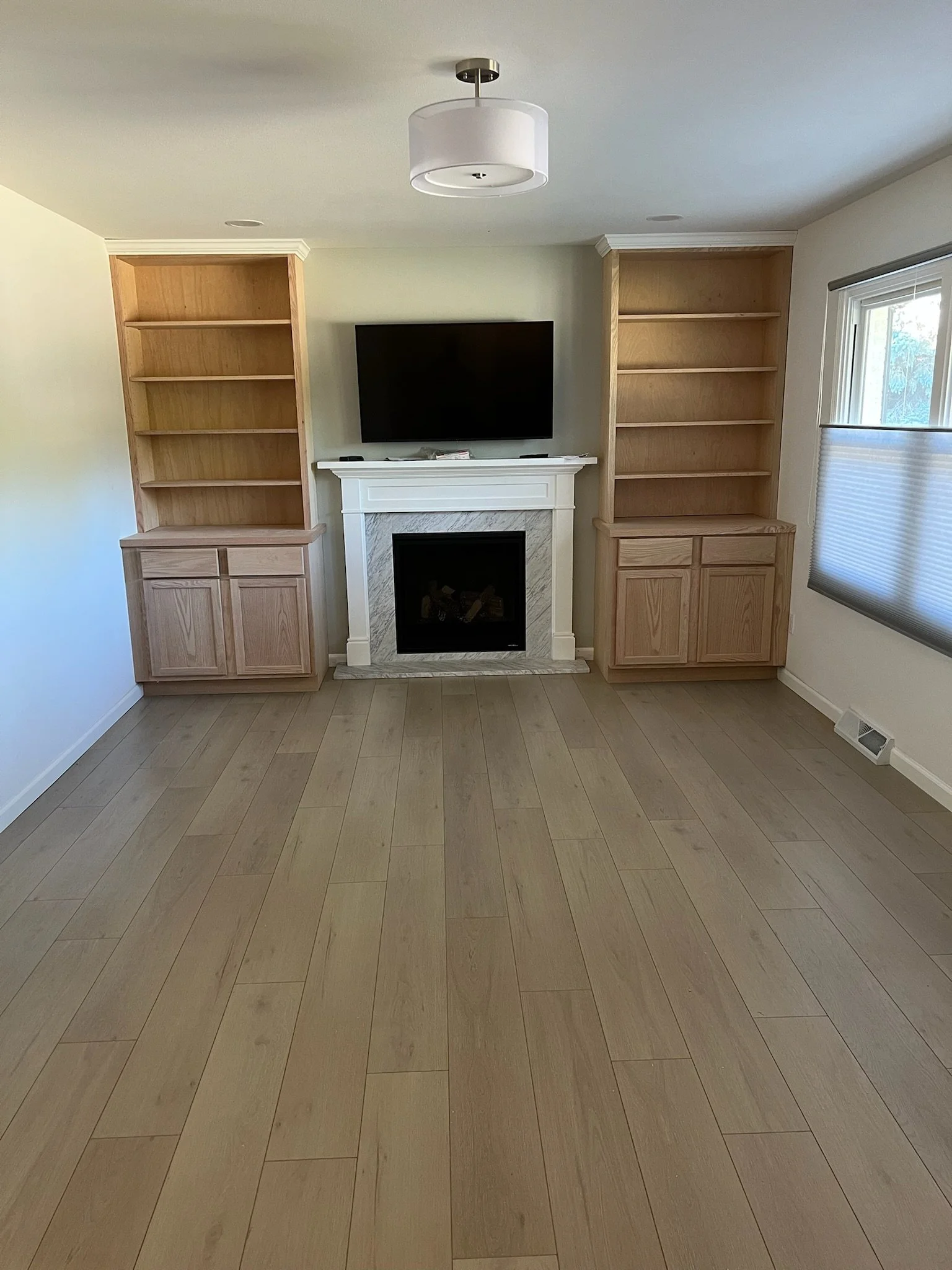 Living room with light-colored wooden flooring, white walls, twin built-in bookshelves flanking a fireplace, a mounted flat-screen TV above the fireplace, a window with blinds on the right, and  ceiling light fixture. Iowa City. Stecker Home Services