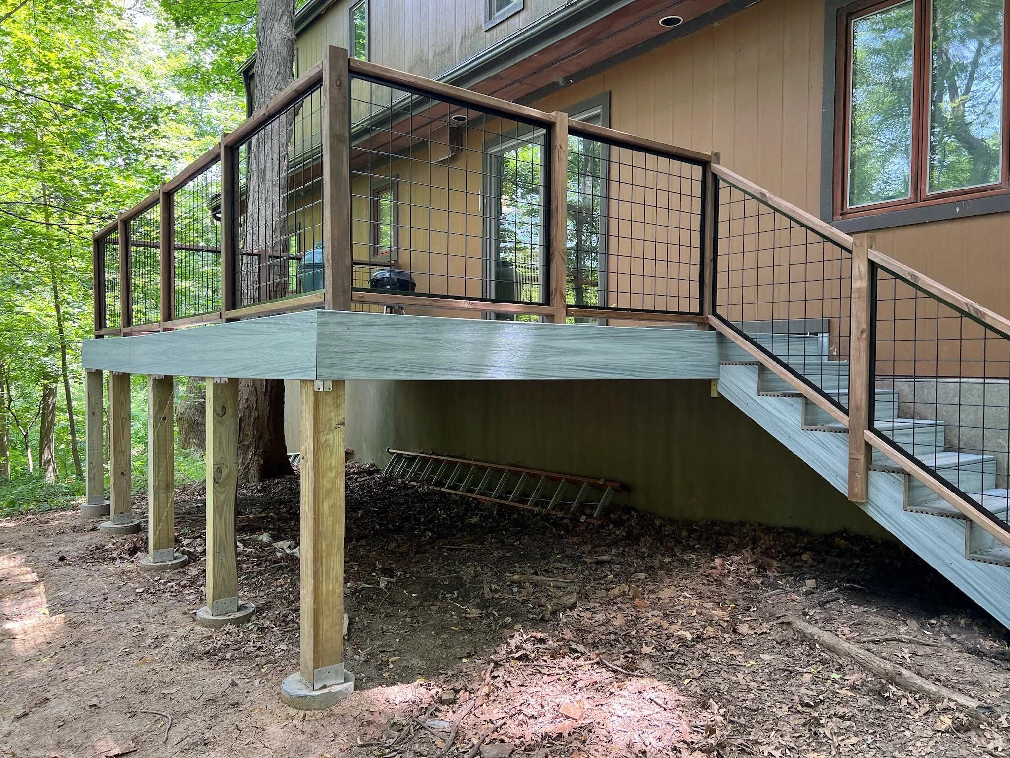 Wooden deck with black metal railing attached to a house with tan siding and large windows, surrounded by trees and dirt ground underneath. Iowa City. Stecker Home Services. Home Renovations. Iowa City Contractor. 