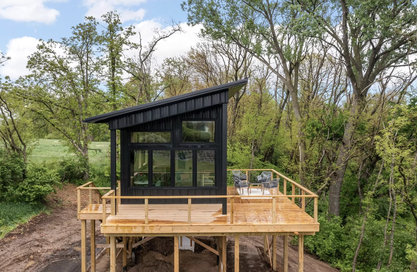 A modern black tiny house with a sloped roof on a raised wooden deck, surrounded by green trees and a blue sky. Iowa City. Stecker Home Services. Home Renovations. Iowa City Contractor. 