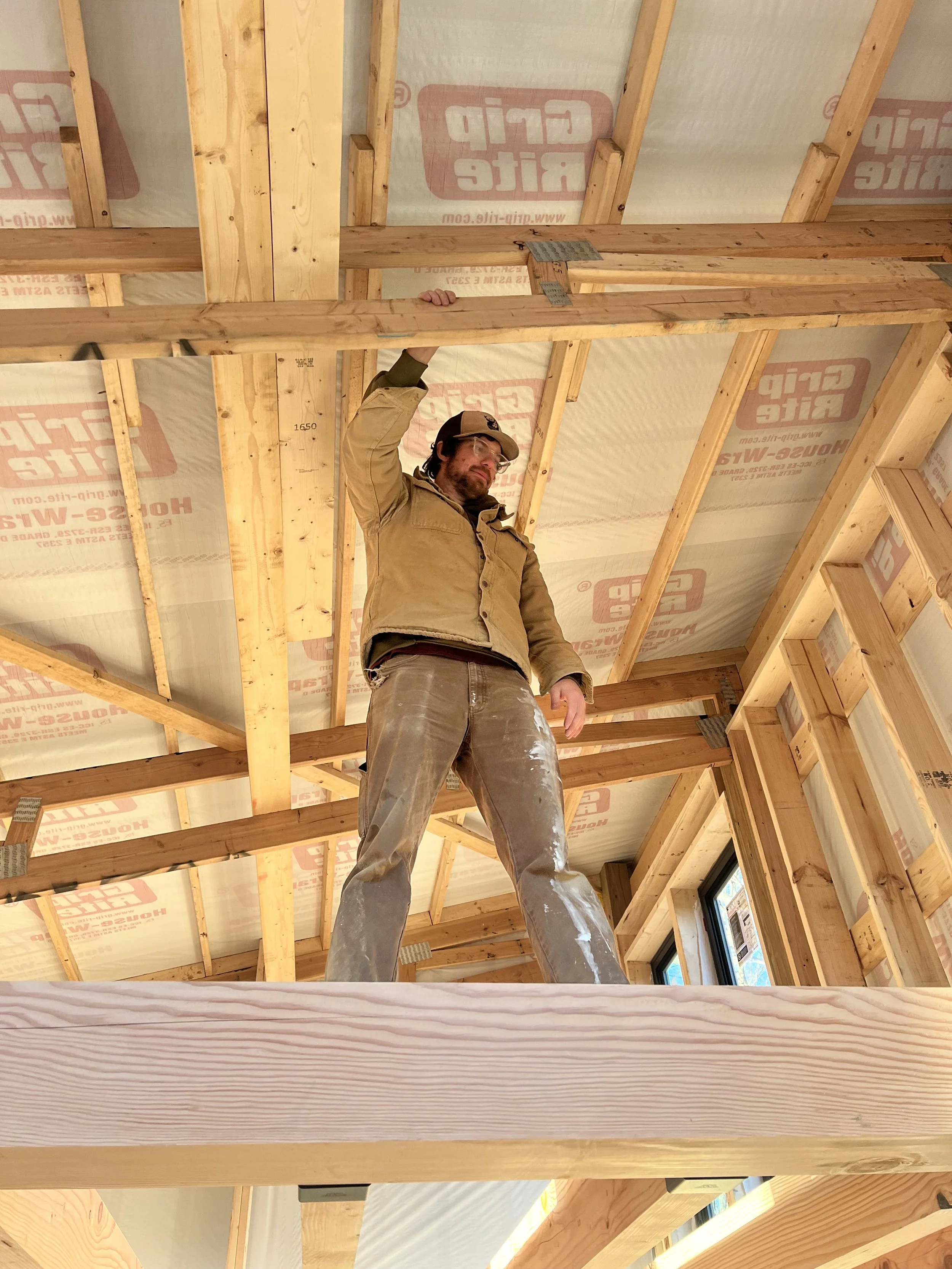 A man with glasses and a baseball cap working on framing in a house under construction, surrounded by wooden beams and construction materials.