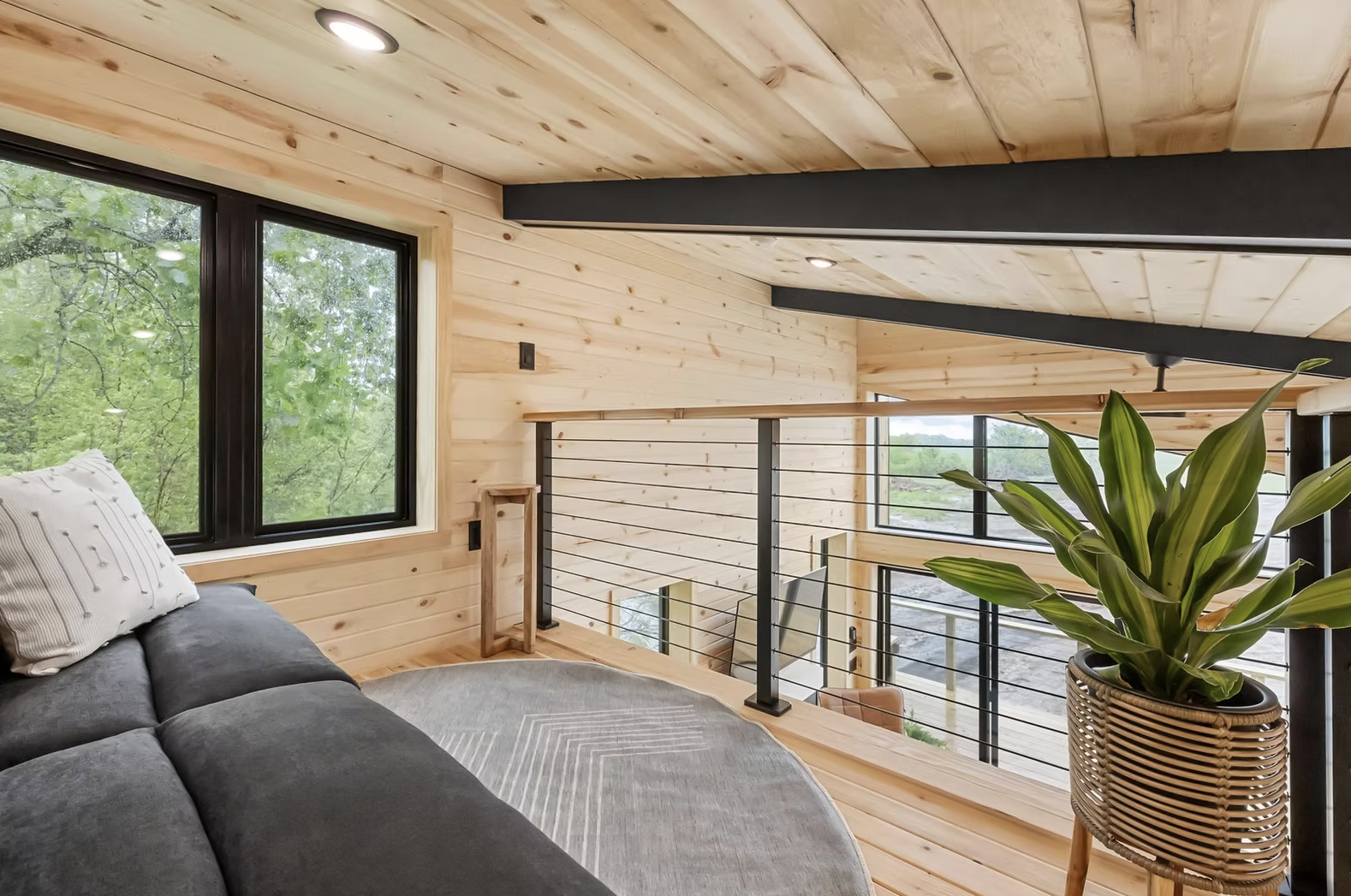 Interior of a modern wooden house with large windows showing green trees outside, a gray couch with a white pillow, a potted plant on a woven stand, and a wooden railing overlooking the lower level. Iowa City. Stecker Home Services. Home Renovations.
