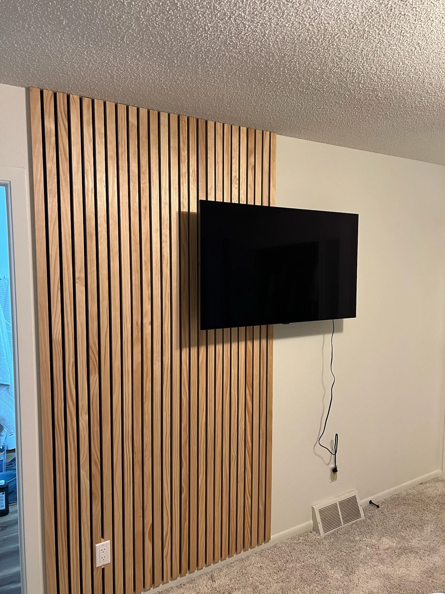 A flat-screen television mounted on a wall next to a decorated wooden wall panel, with an electrical outlet and vent nearby, and a power cord hanging down. Iowa City. Stecker Home Services. Home Renovations. Iowa City Contractor. 