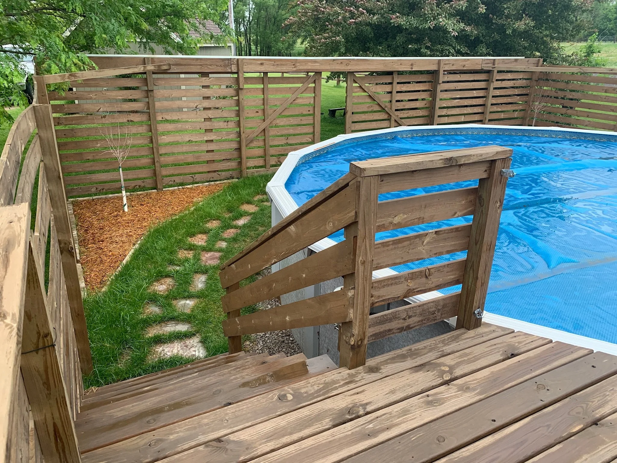 An above view of a backyard pool area enclosed by a wooden fence with a small tree beside the fence, and a grassy lawn with a pathway of stepping stones leading to the pool. Iowa City. Stecker Home Services. Home Renovations. 