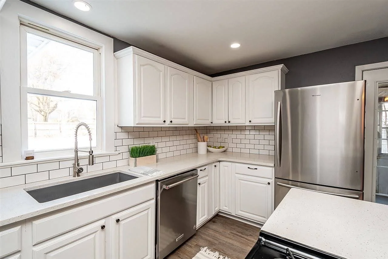 Modern kitchen with white cabinets, a stainless steel refrigerator, a white countertop, and a white subway tile backsplash. A window above the sink lets in natural light. Iowa City. Stecker Home Services. Home Renovations. 