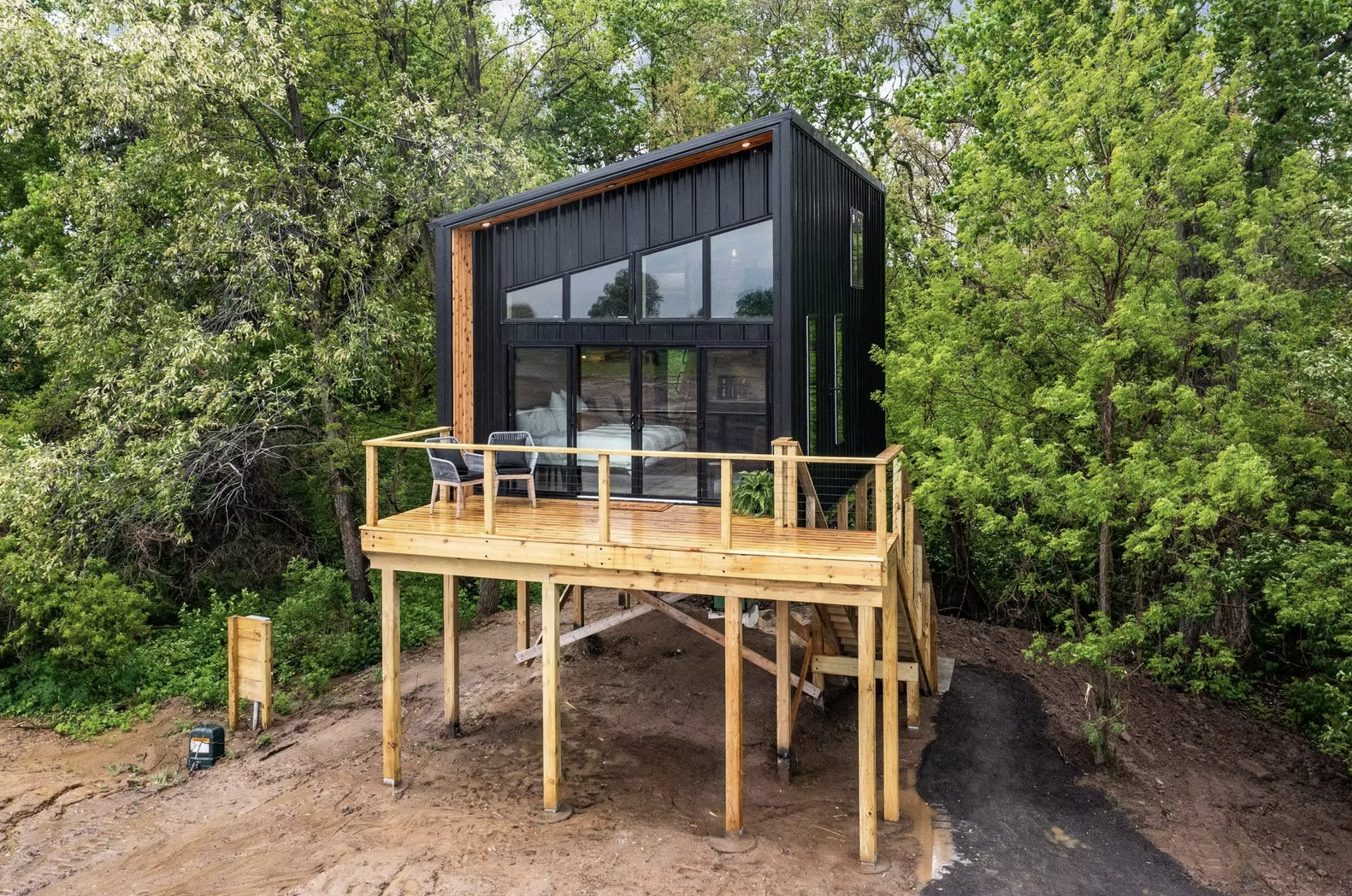 A modern tiny house with black exterior walls, large windows, and a wooden deck with two chairs, situated among green trees on a sloped dirt lot. Iowa City. Stecker Home Services. Home Renovations.