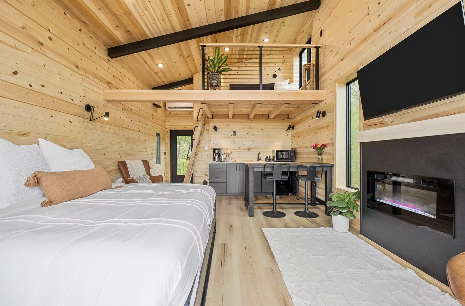 A cozy tiny house interior with wood-paneled walls, a bed, a kitchenette, a small loft area, and a fireplace. Iowa City. Stecker Home Services. Home Renovations. 