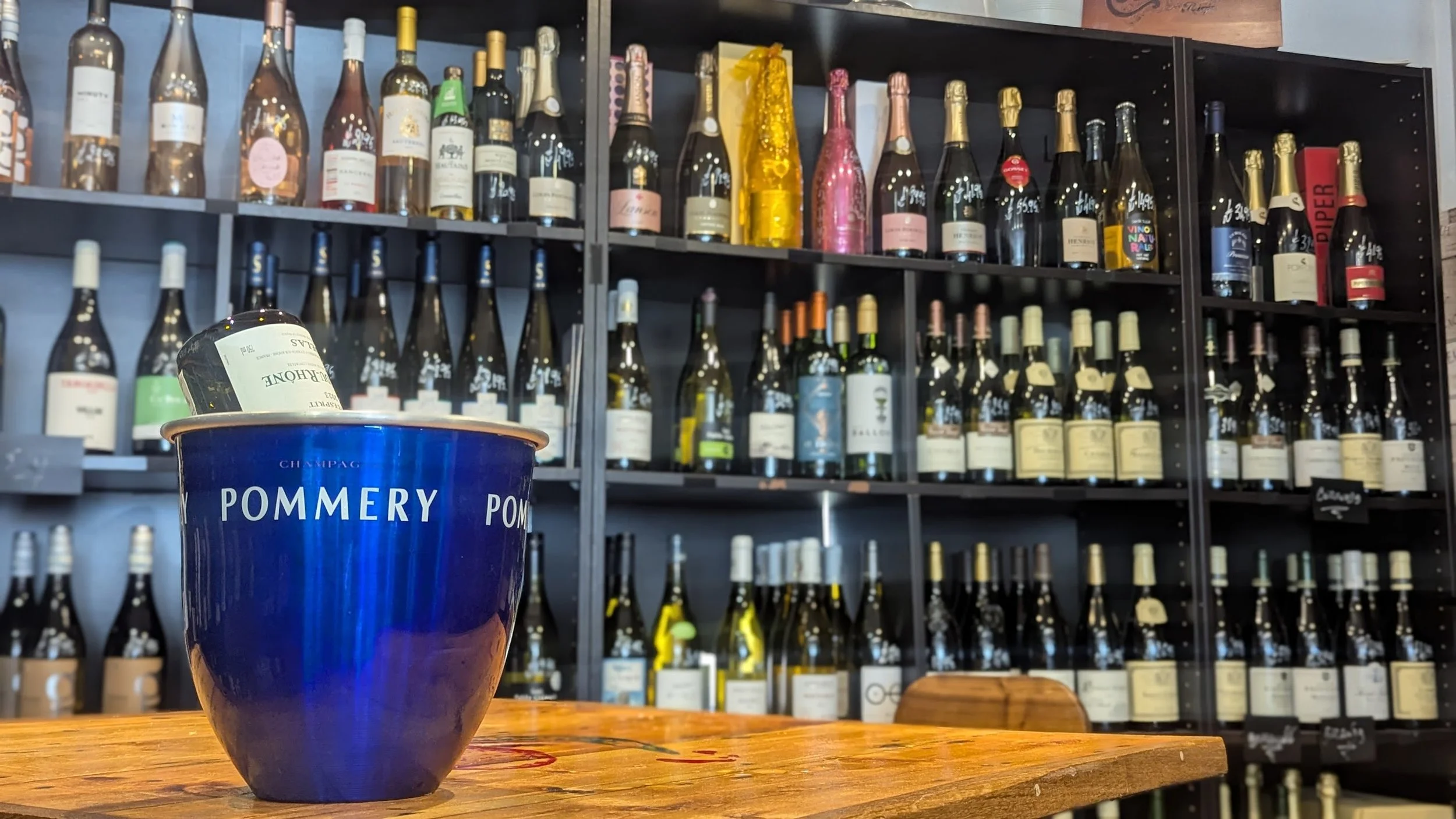 A blue cup labeled 'POMMERY' on a wooden table in front of a wine rack filled with bottles of wine, champagne, and sparkling wine.