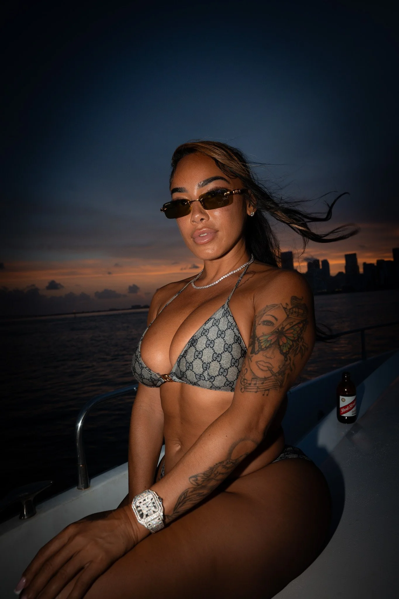 A woman with tattoos and gold-rimmed sunglasses sitting on a boat at sunset, wearing a patterned bikini top and jewelry.