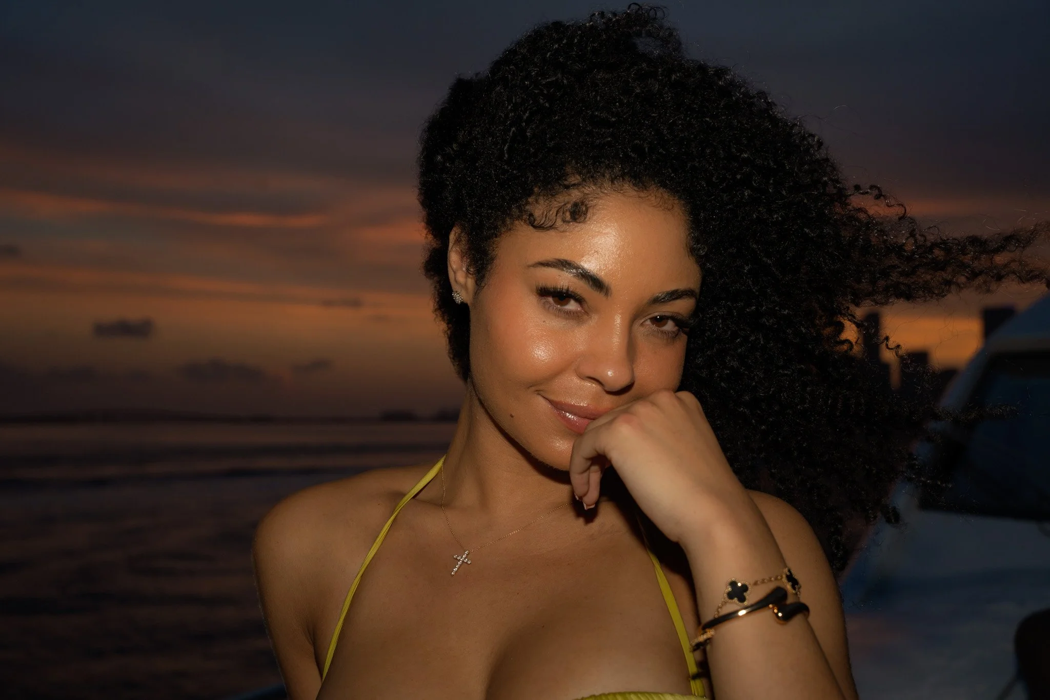 A woman with curly dark hair and light skin smiling at sunset on the beach, wearing a yellow top and jewelry.