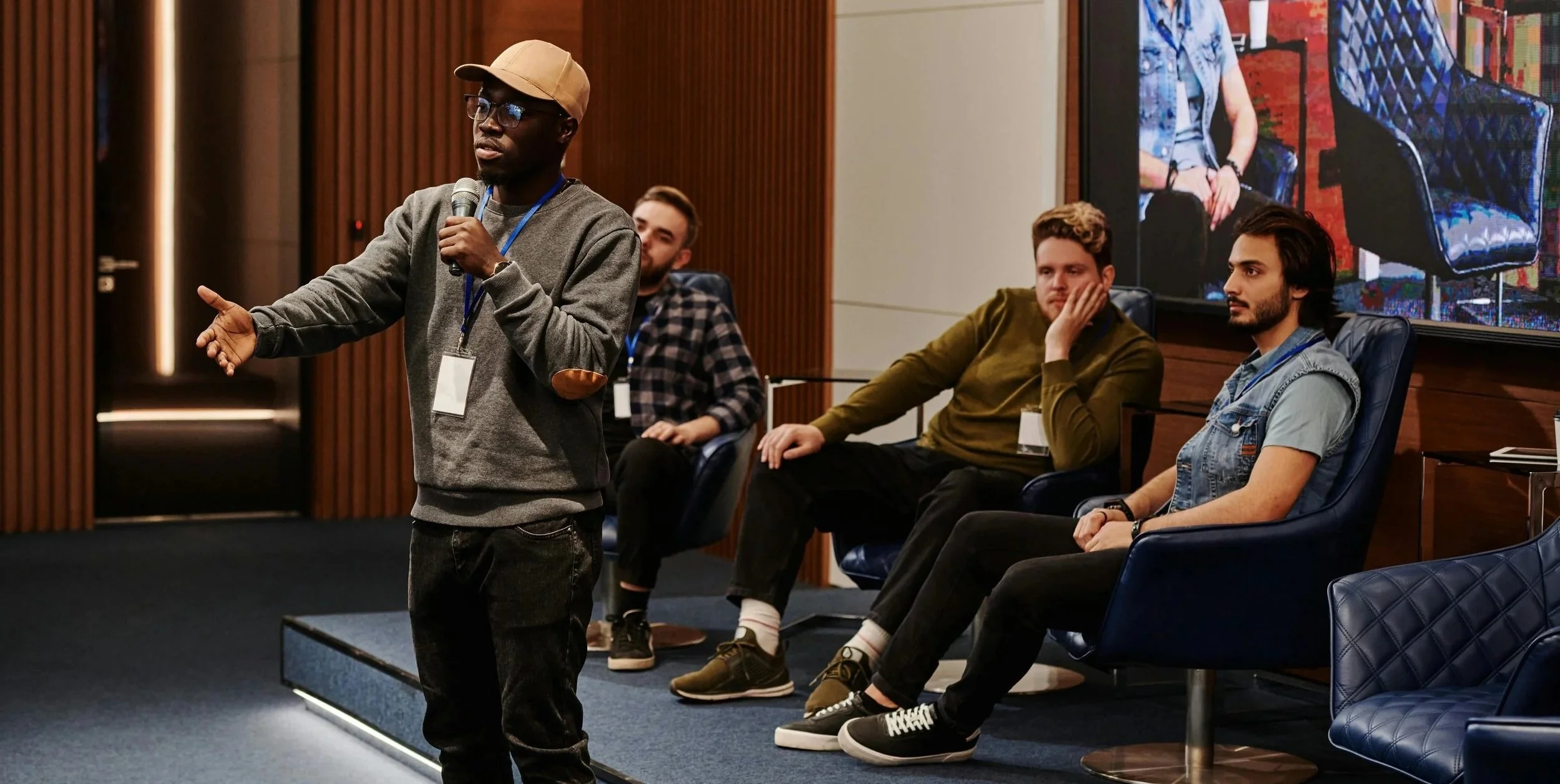 A man in a gray sweatshirt and beige cap holding a microphone and speaking at a panel discussion, with three men seated in chairs listening.